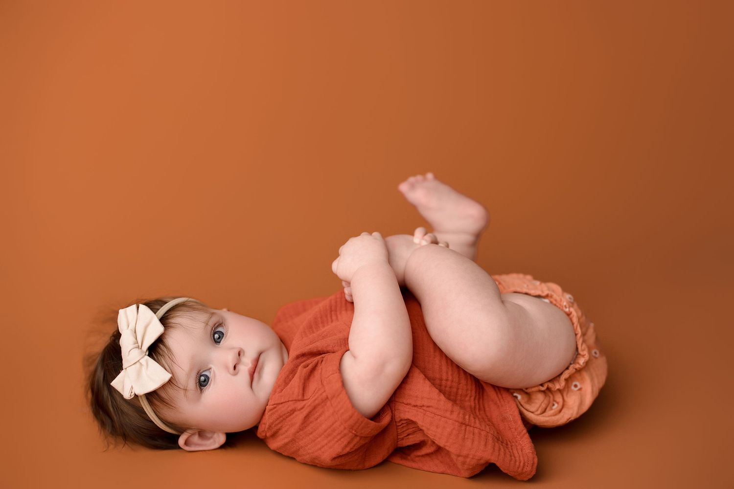 Newborn baby wearing coral colored outfit lies on warm brown backdrop in artistic studio portrait series.