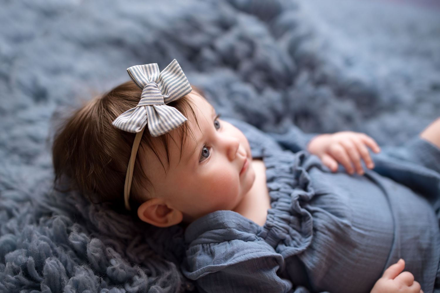 A young child wearing a white bow lies on a gray blanket while looking upward with a peaceful expression.