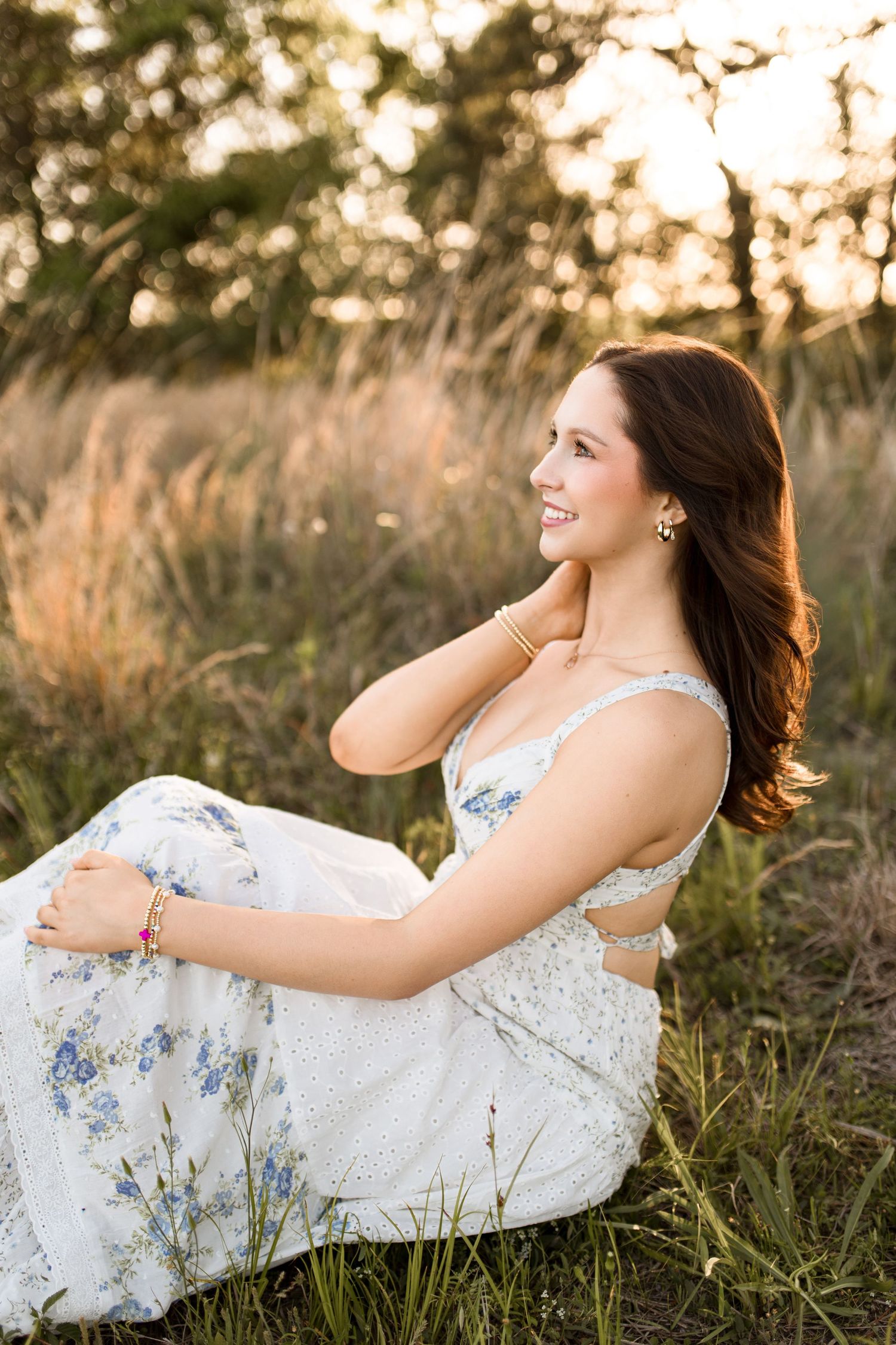 Young woman in white floral dress sitting in golden field at sunset, smiling and touching her hair.