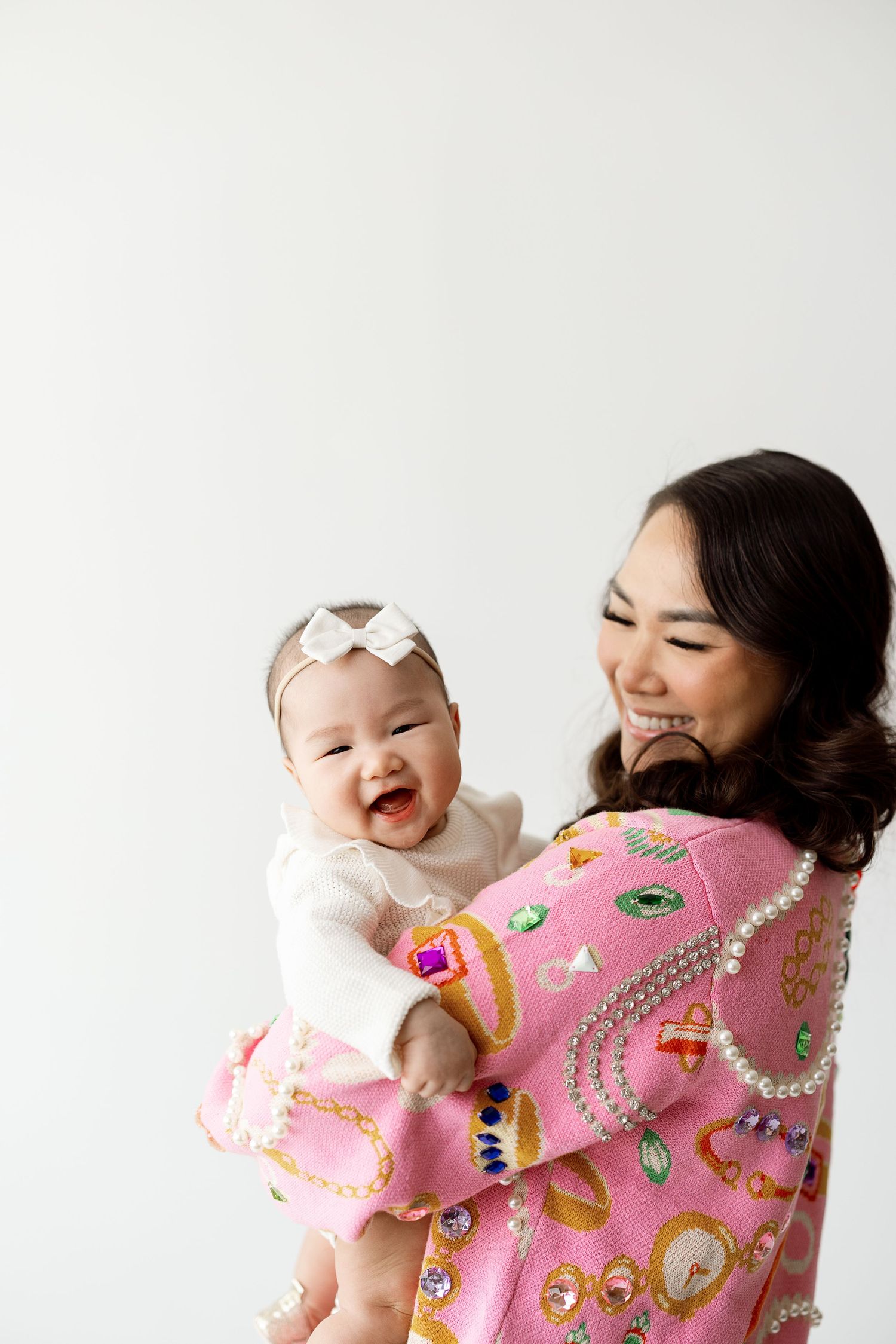 Joyful interaction between mother wearing pink kimono and baby in white outfit on light background.