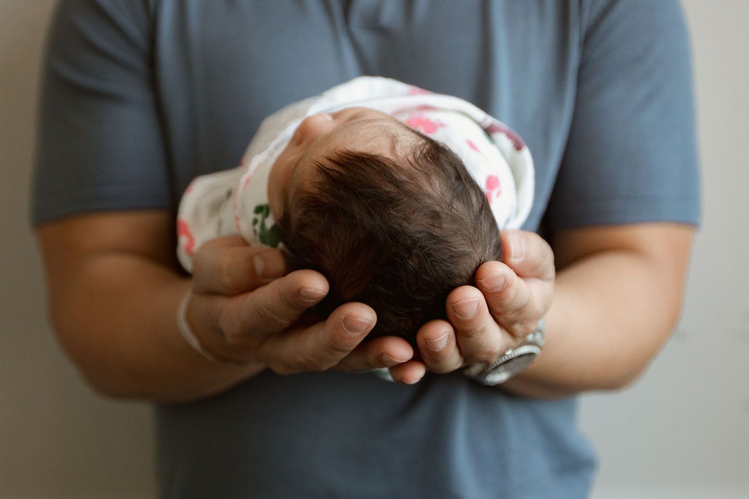 Gentle hands cradling newborn wrapped in patterned white blanket.