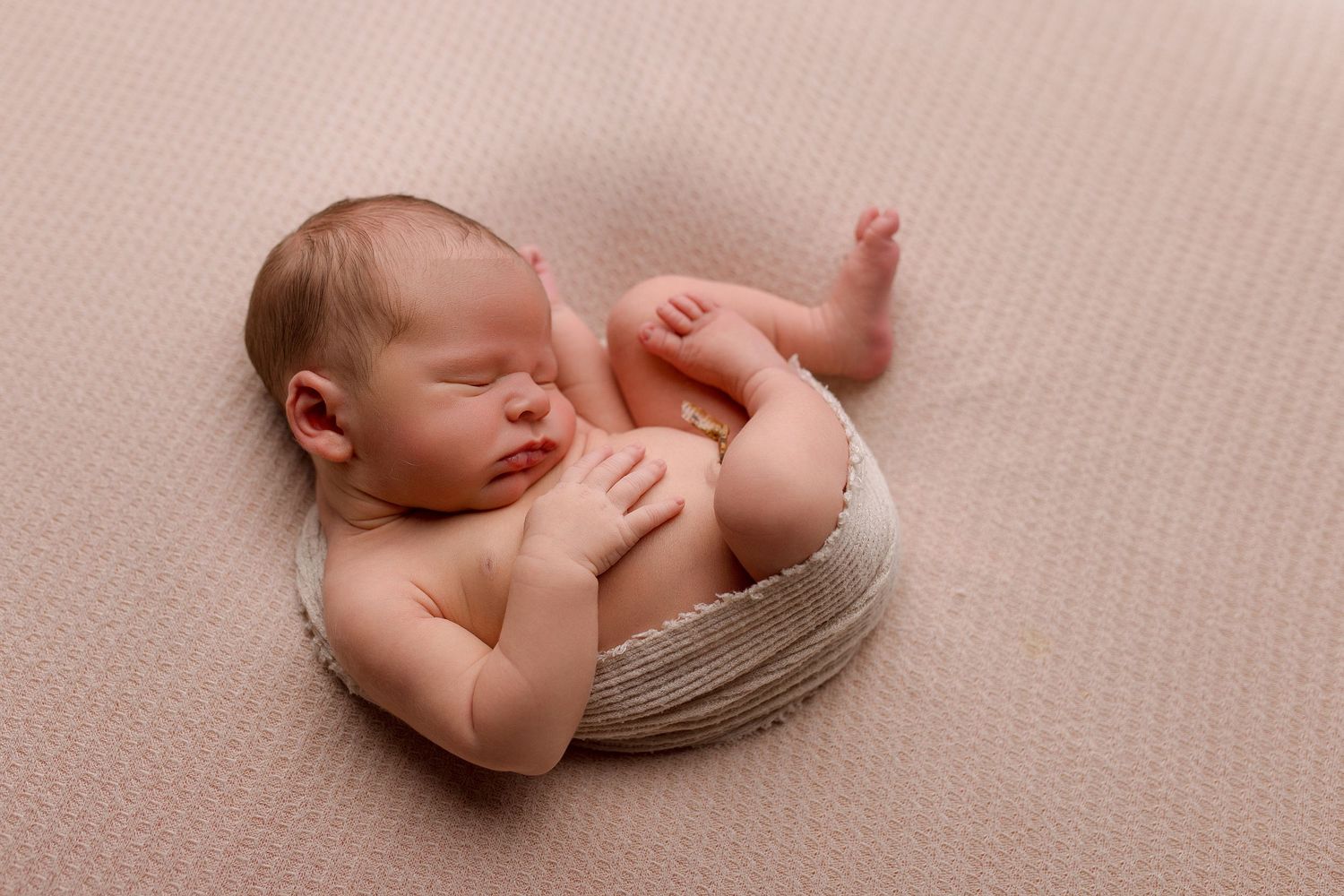 Peaceful newborn baby sleeping curled up in a bowl wrap on a soft pink blanket in a studio photo.