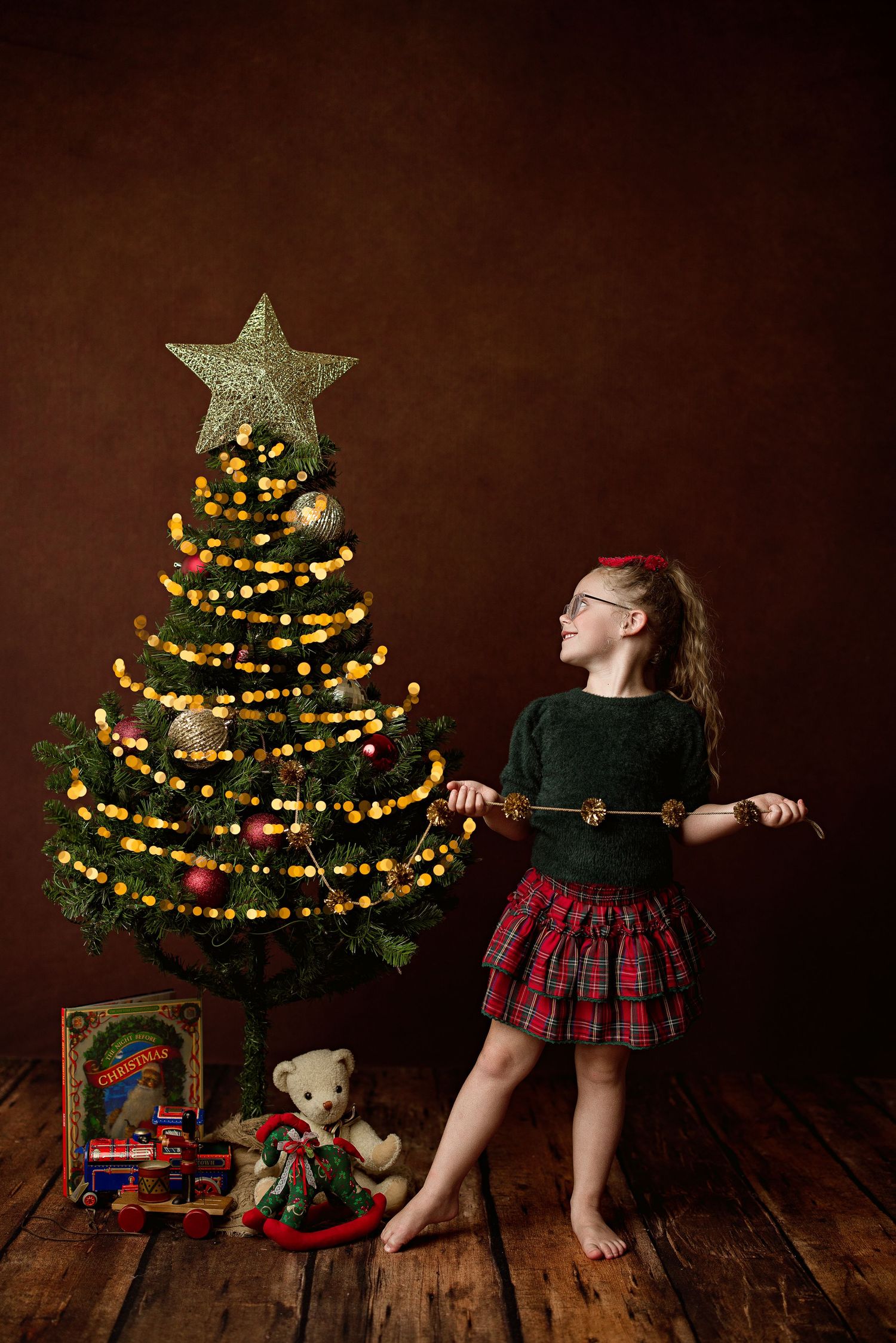Creative Christmas photo series shows someone pulling a decorated tree across wooden floor with wrapped presents underneath.