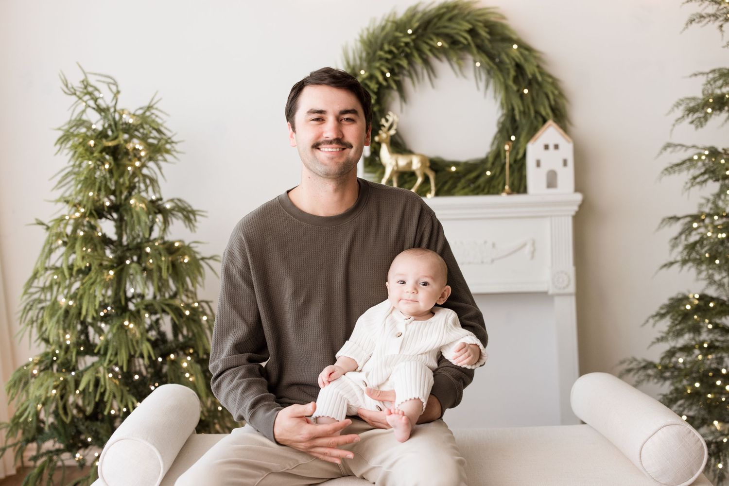 A parent holds a baby while sitting on a white couch in a cozy living room with green plants.