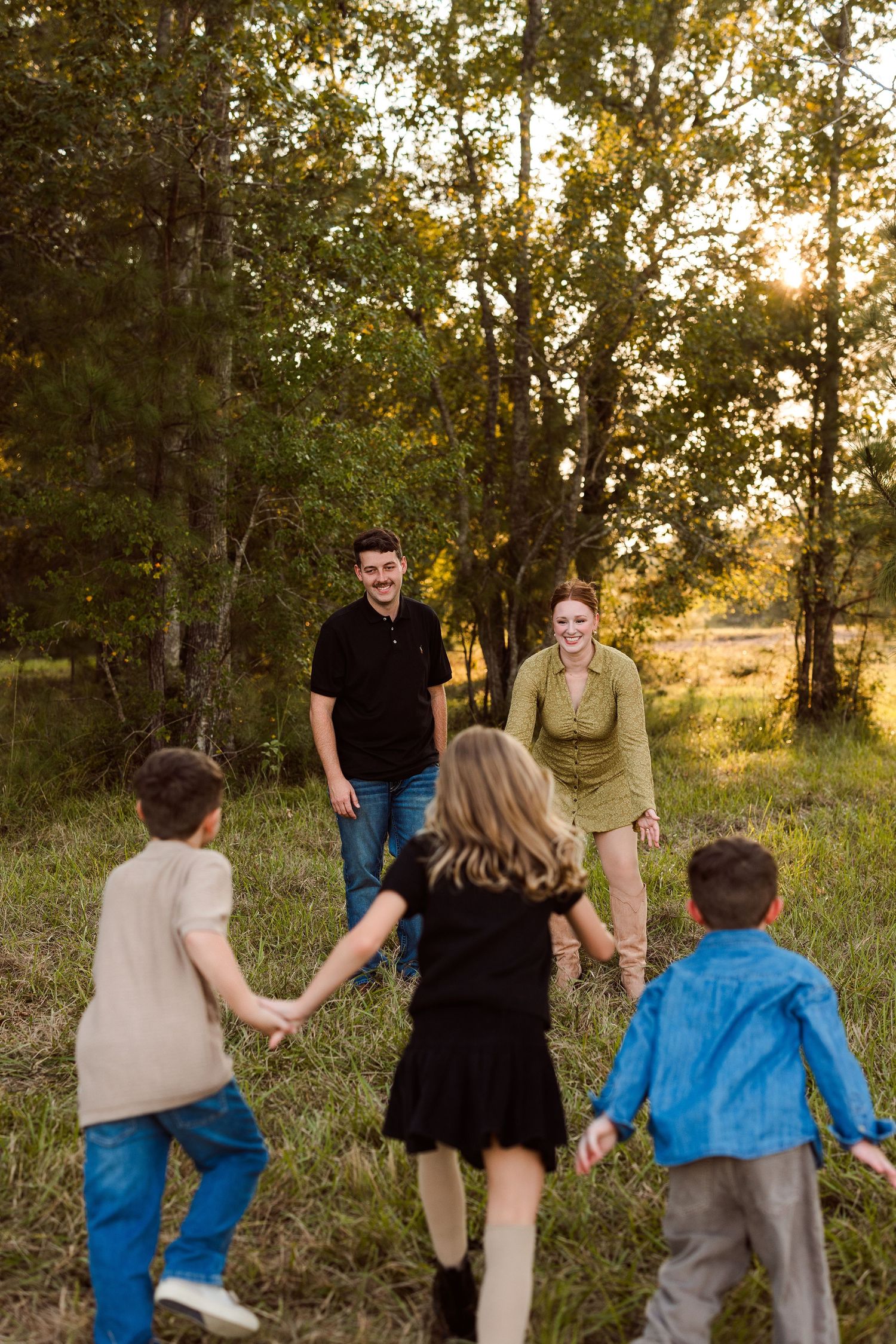 Three children hold hands and run across a sunny grassy field toward smiling parents standing near tall trees during a family photo session.