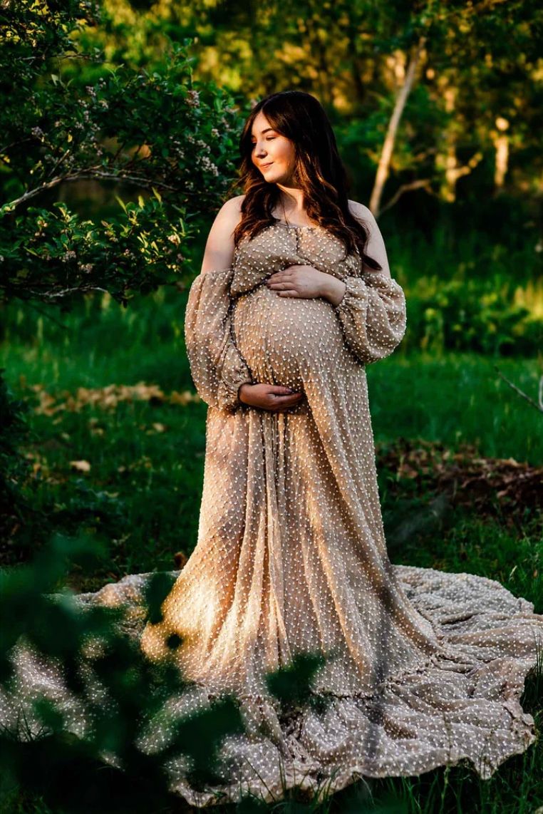 An expectant woman stands in a flowy lace dress with long sleeves in a sunlit garden setting during a maternity photoshoot.