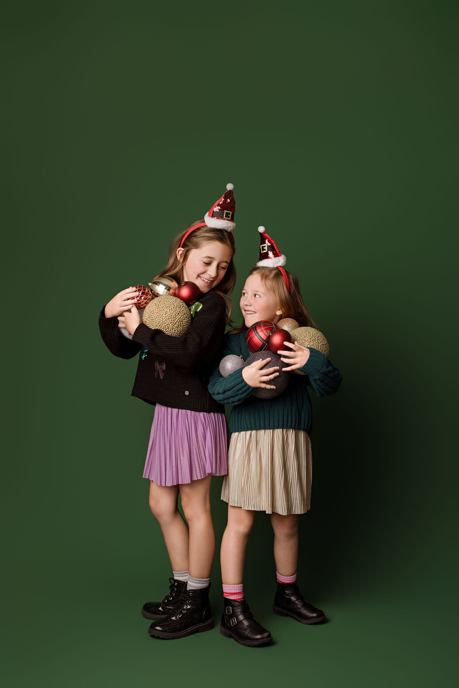Two young children in pleated skirts and dark shoes pose playfully against a dark green studio backdrop.