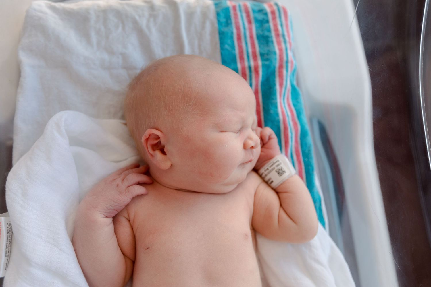 A peaceful sleeping newborn baby rests on white blankets with a striped blue and teal blanket nearby during Fresh 48 session in Beaumont.