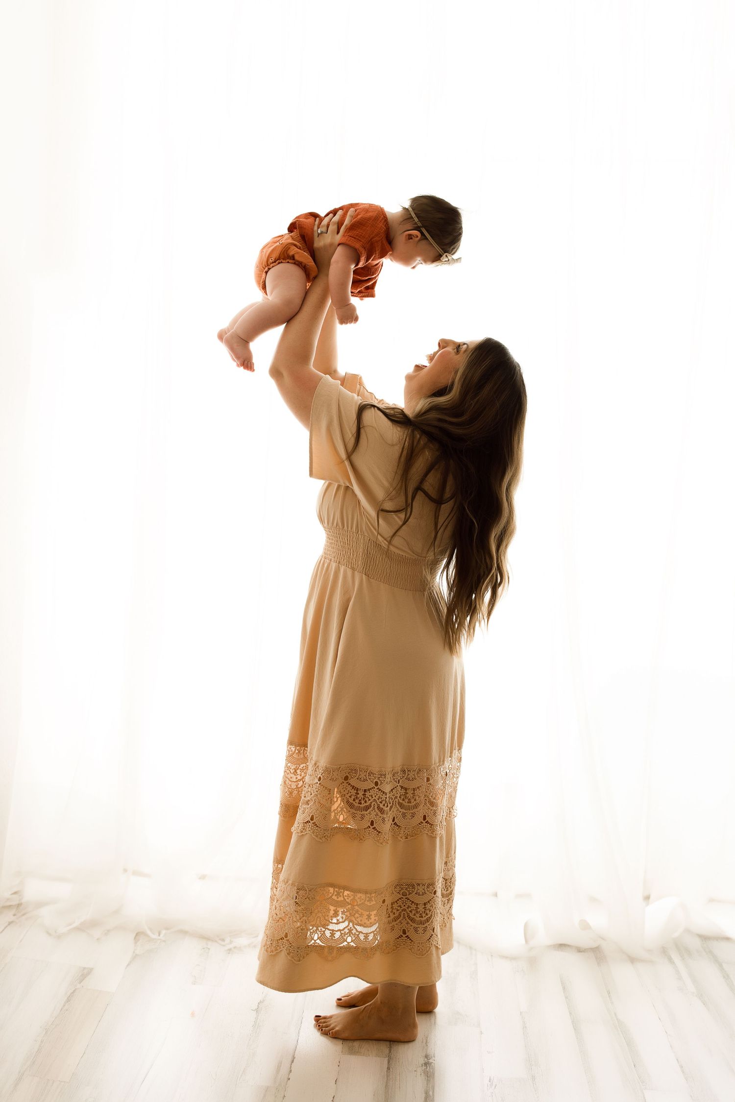 Woman in beige dress holds baby tenderly in bright white studio setting, creating intimate mother-child moments.