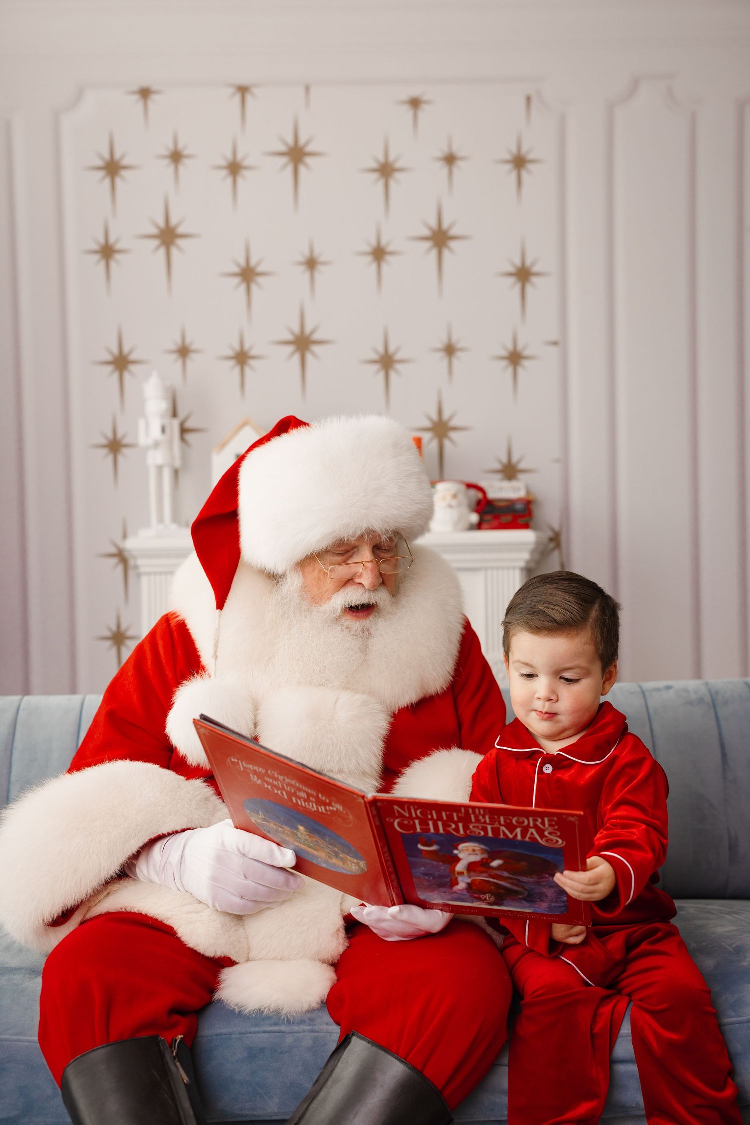 Santa Claus reads a Christmas storybook to a young child wearing red on a gray couch with star decorations behind them.