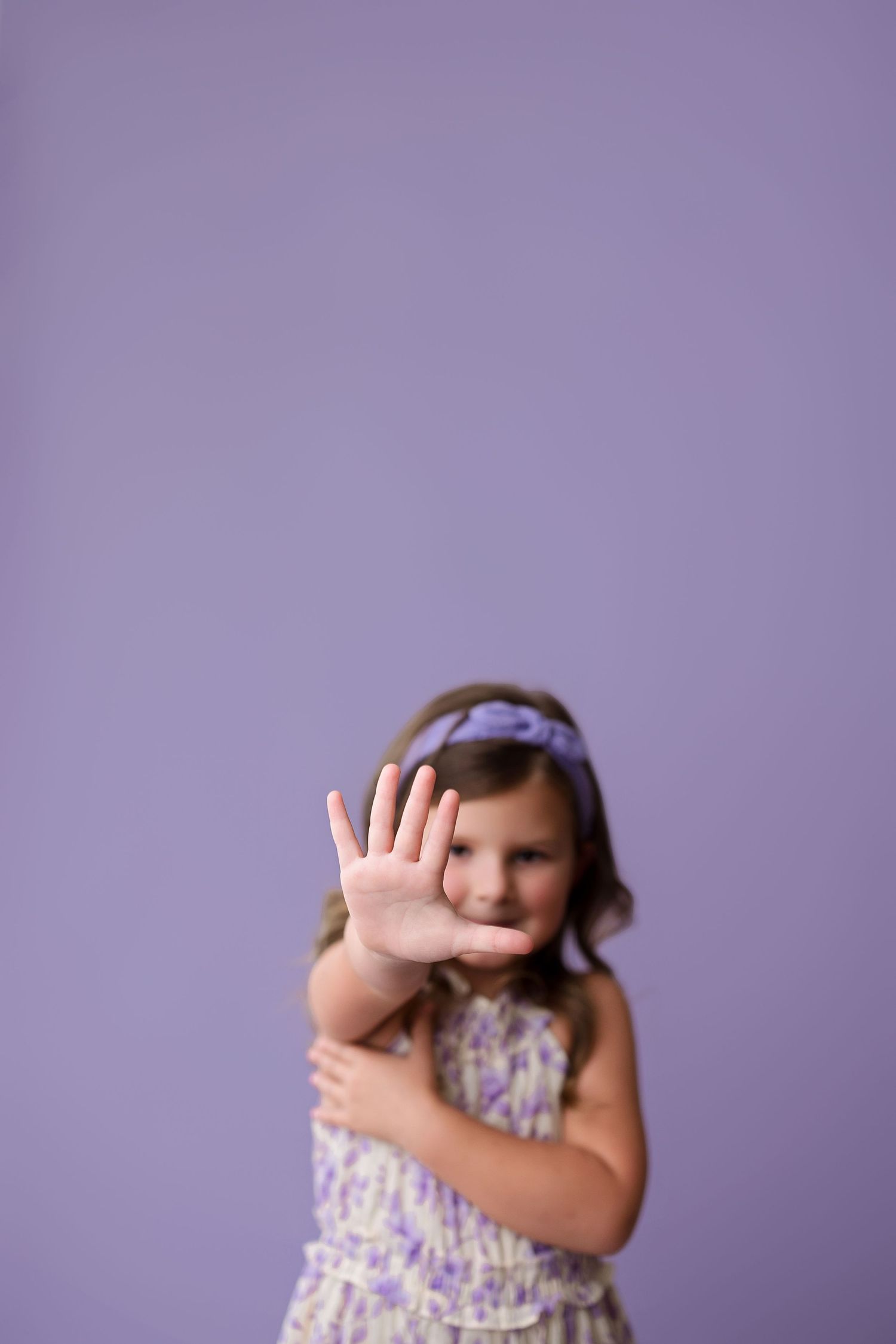 Child wearing floral dress making stop gesture with hand against purple wall.