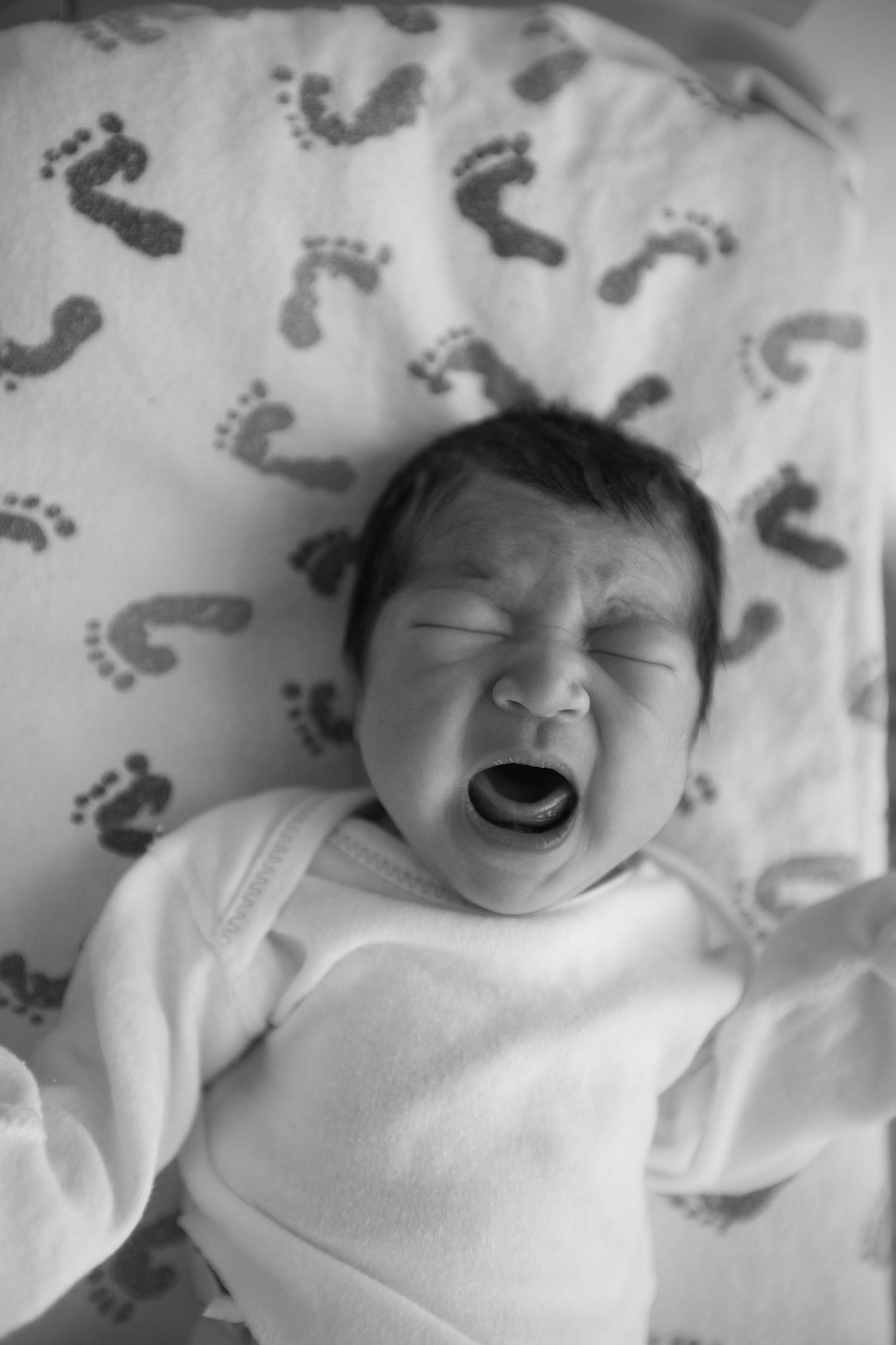 A black and white portrait of a crying newborn on a footprint-patterned blanket.