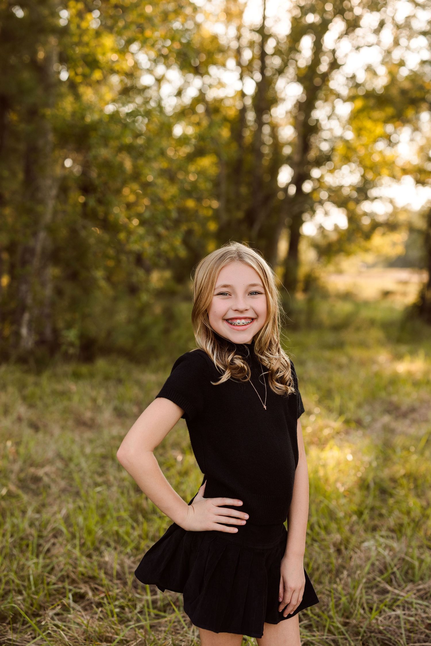 Smiling girl with long wavy blonde hair in a black short-sleeve dress poses with a hand on her hip in a sunlit grassy field with trees.