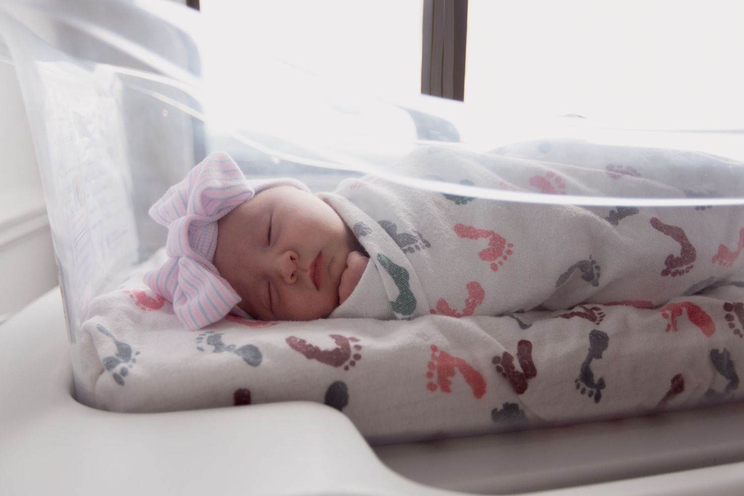Newborn baby sleeping peacefully in white hospital bassinet with pink and gray patterned blanket.