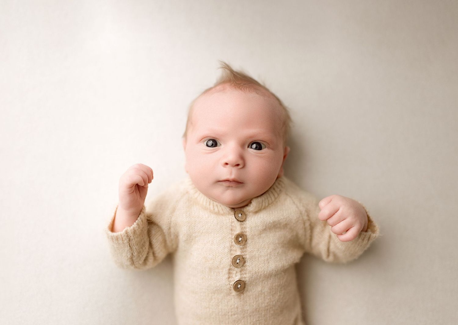 Baby wearing a cozy cream-colored knit sweater lies on white backdrop during professional photo session.