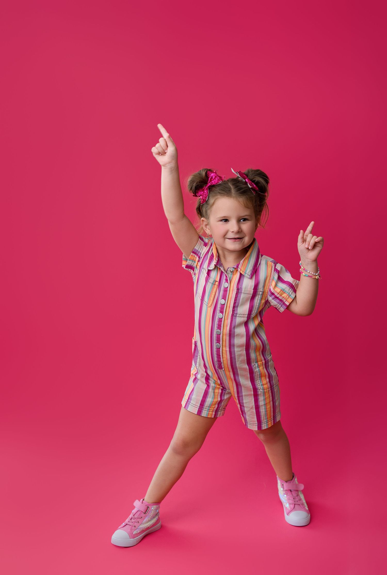 Energetic young child in striped dress and pigtails dances playfully against bright pink background.