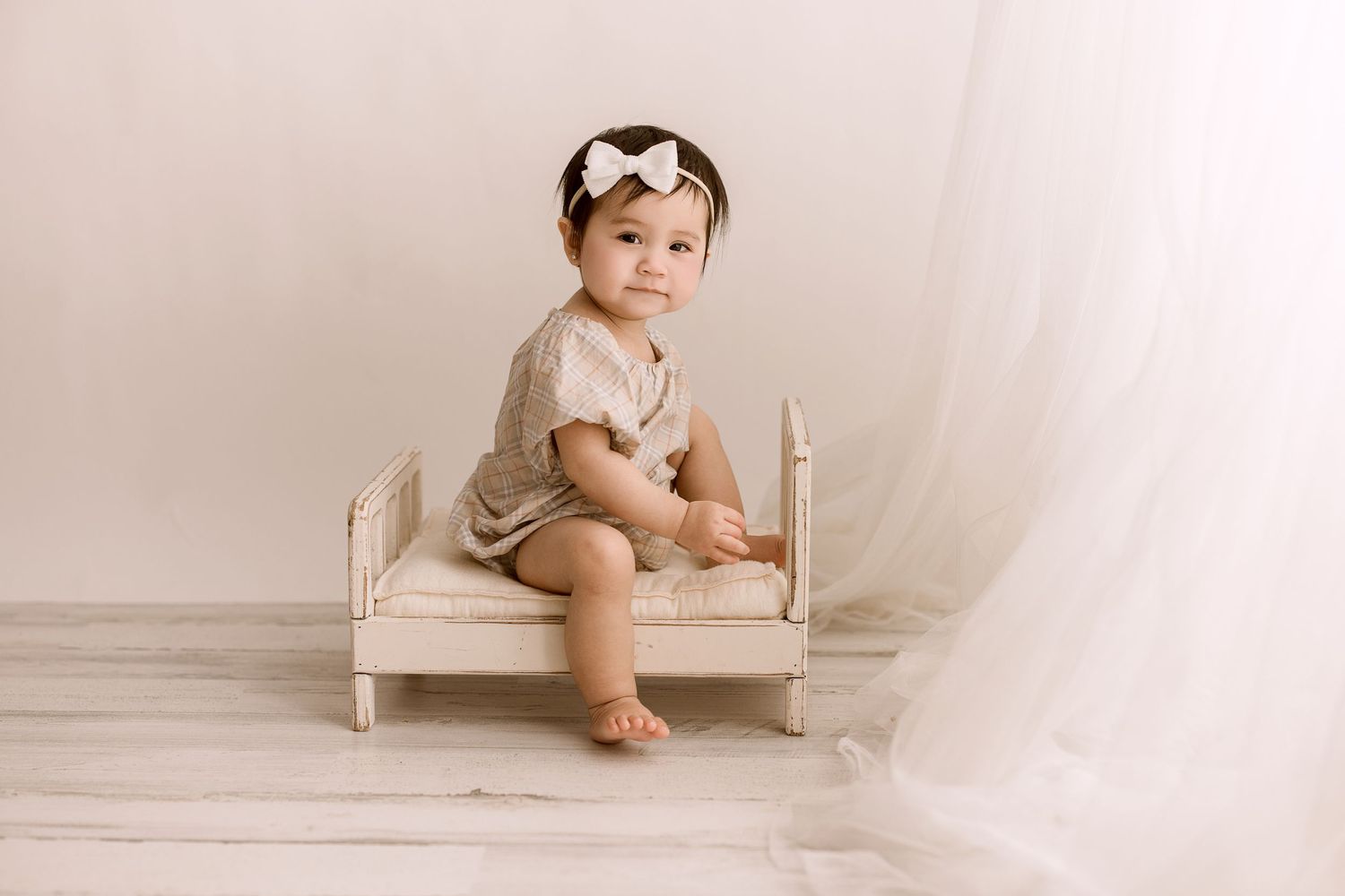 Small child sits on miniature wooden bed wearing cream colored romper against neutral backdrop in studio setting.