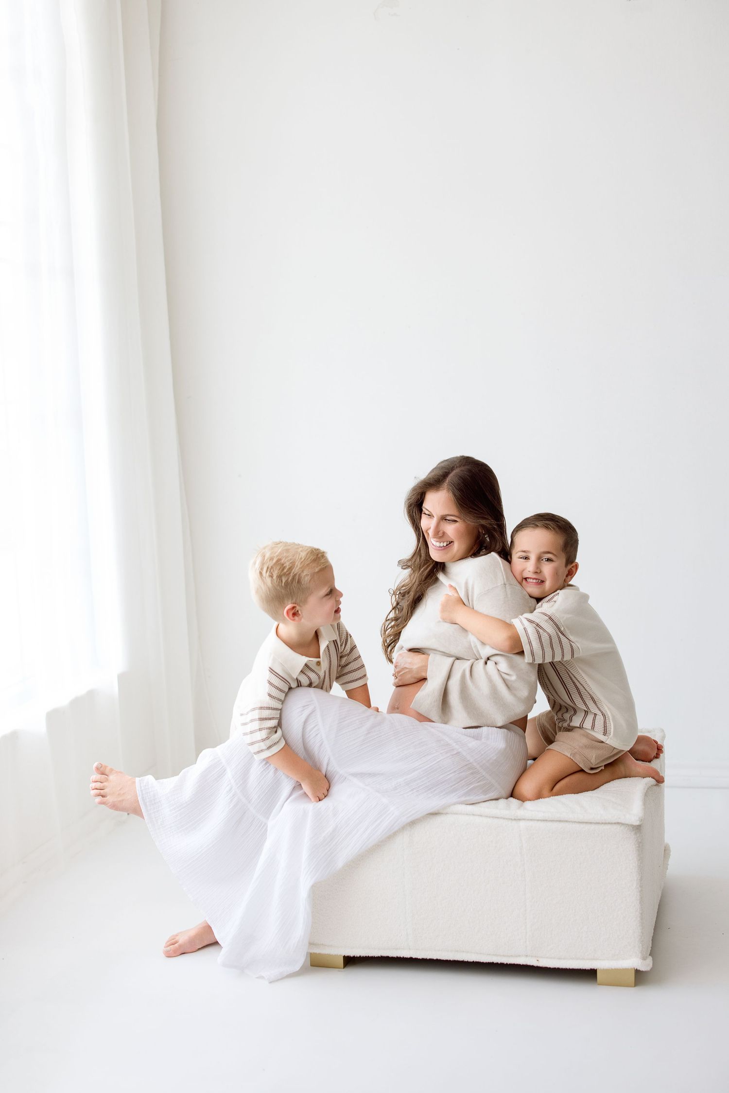 Family wearing neutral colors sits together on white ottoman in bright minimal studio setting with Bree Elle Photography in Beaumont, TX.