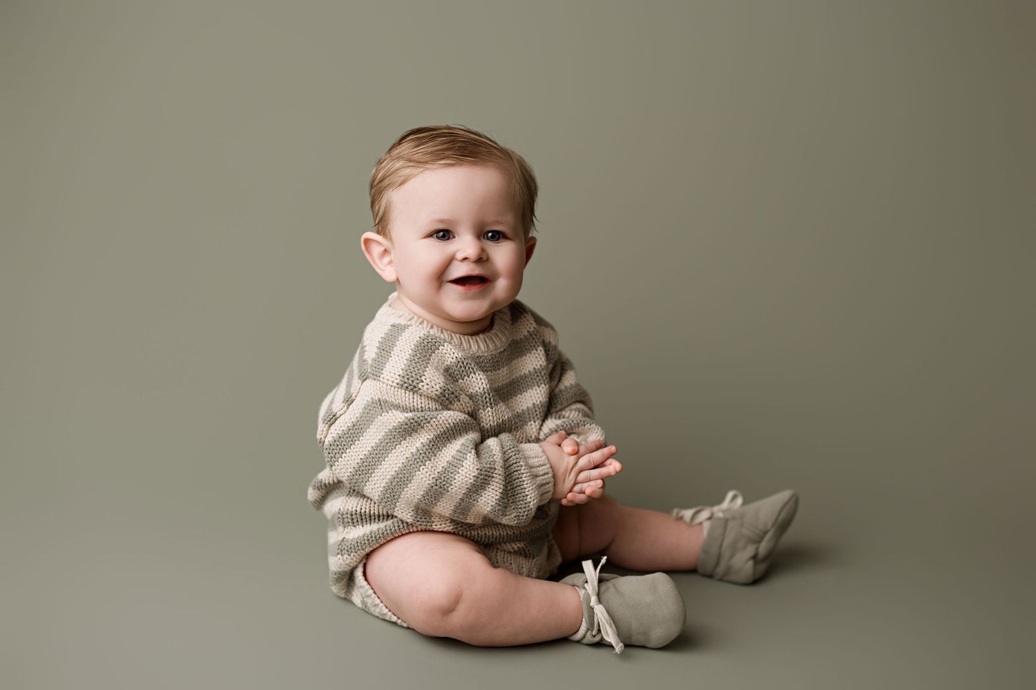 A baby in striped onesie sits on beige backdrop while holding a stuffed bunny toy.