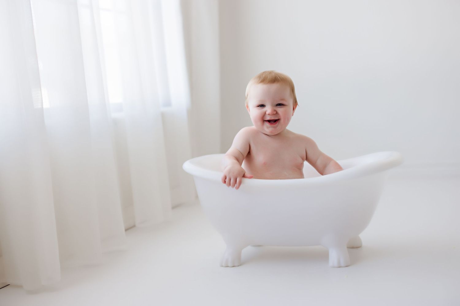 Laughing baby in white clawfoot bathtub against light backdrop.