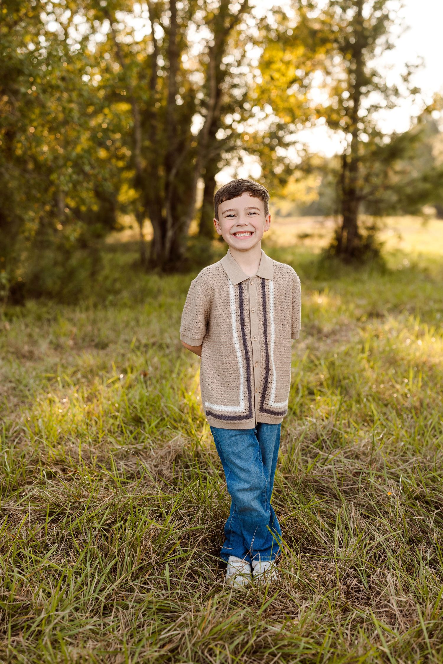 Smiling young boy in a tan striped knit polo shirt and blue jeans stands in a sunlit grassy field with trees in the background.