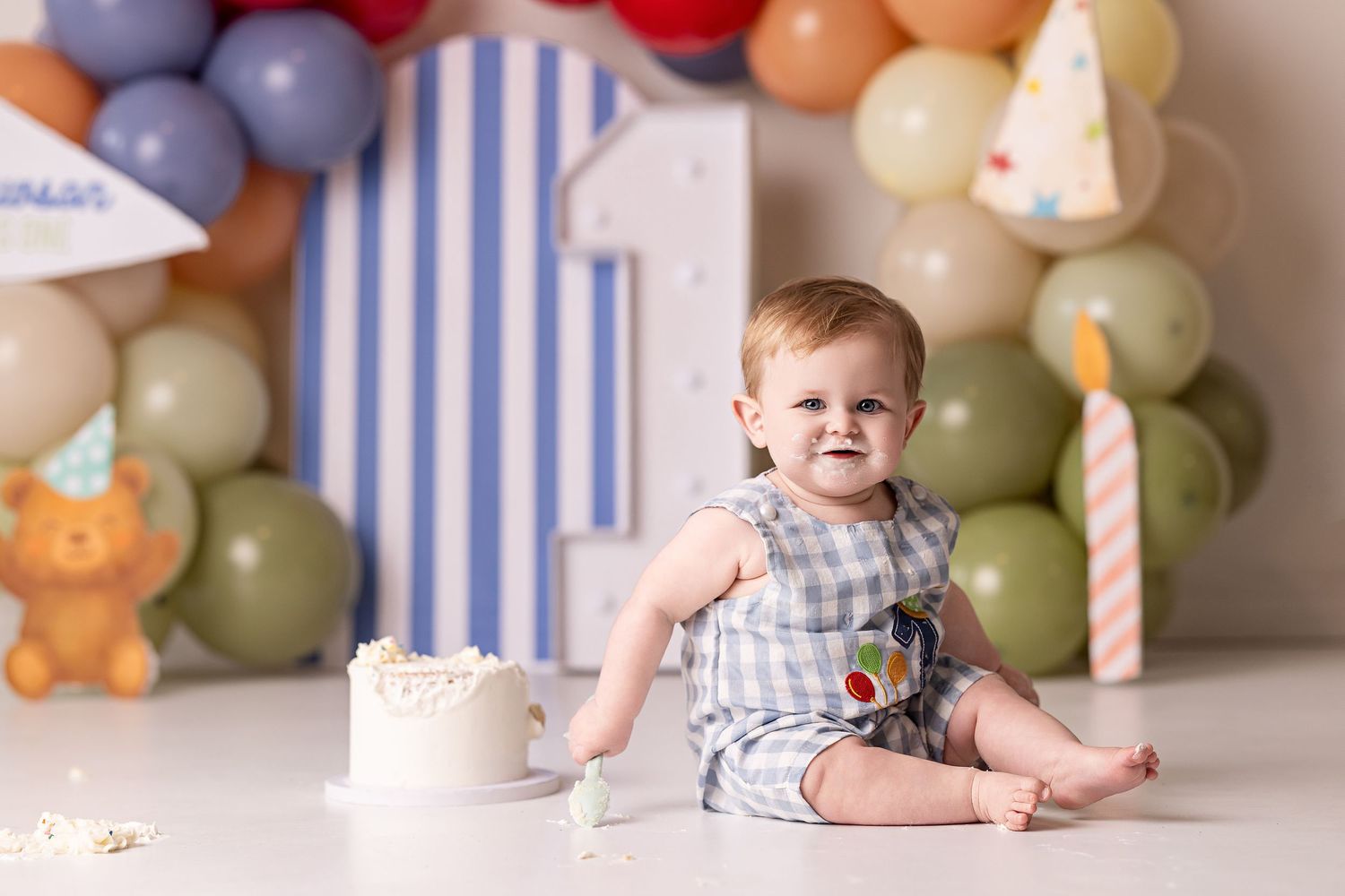 Baby in grey romper sitting beside white birthday cake with festive balloon arch and party decorations.