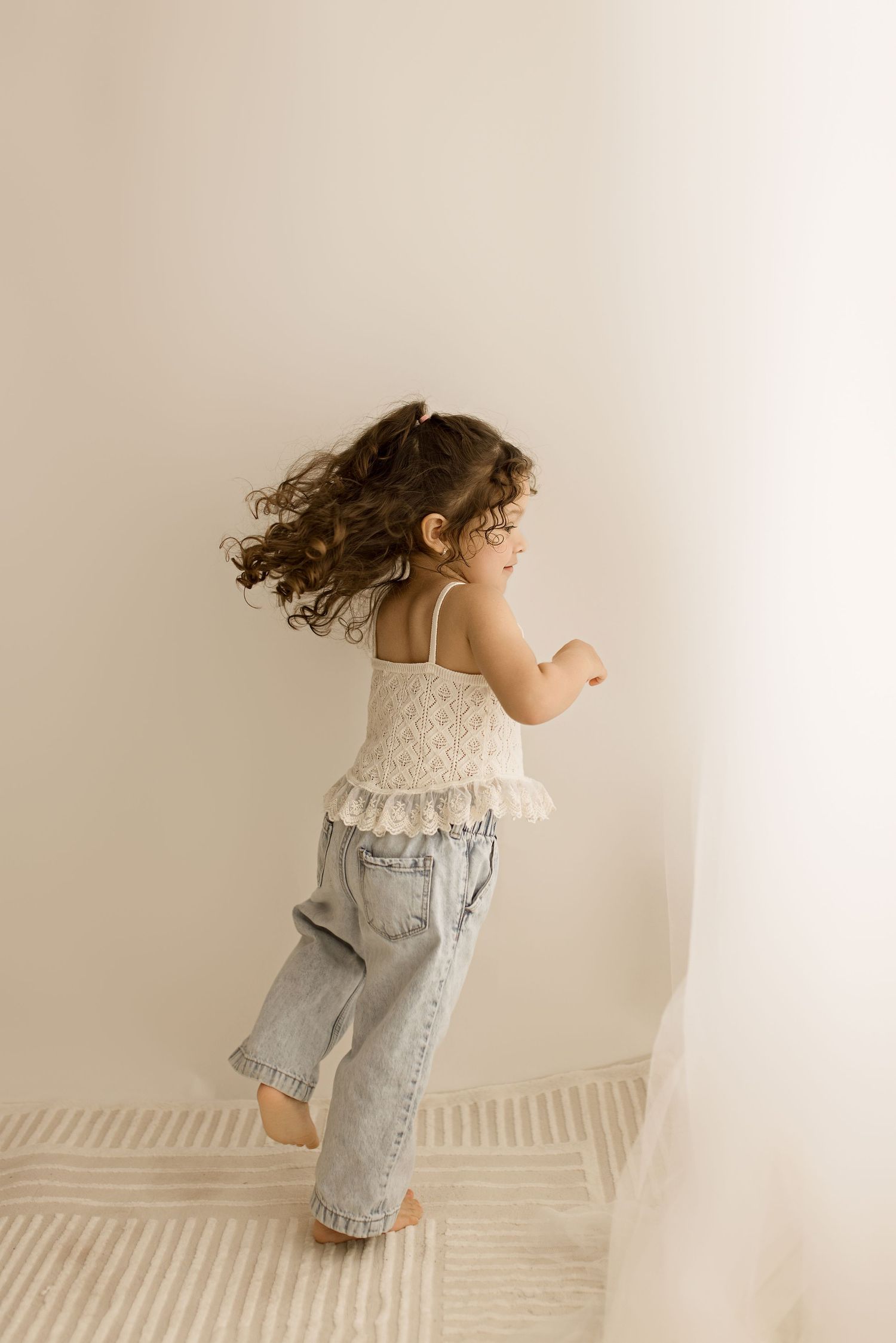 A child in a white lace top and jeans dances joyfully against a neutral wall, their curly hair flowing with movement.