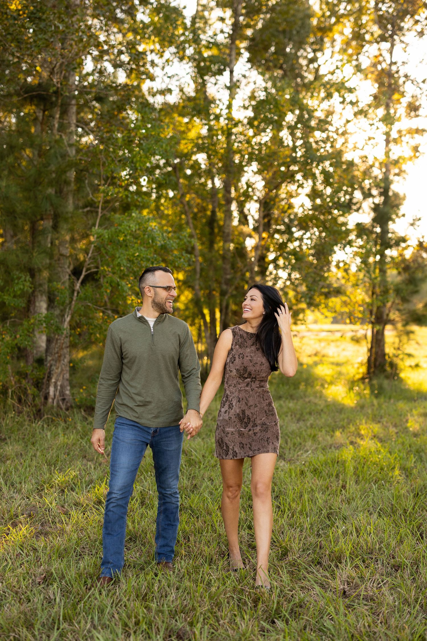 A couple walks hand in hand through a sunlit wooded area during golden hour.