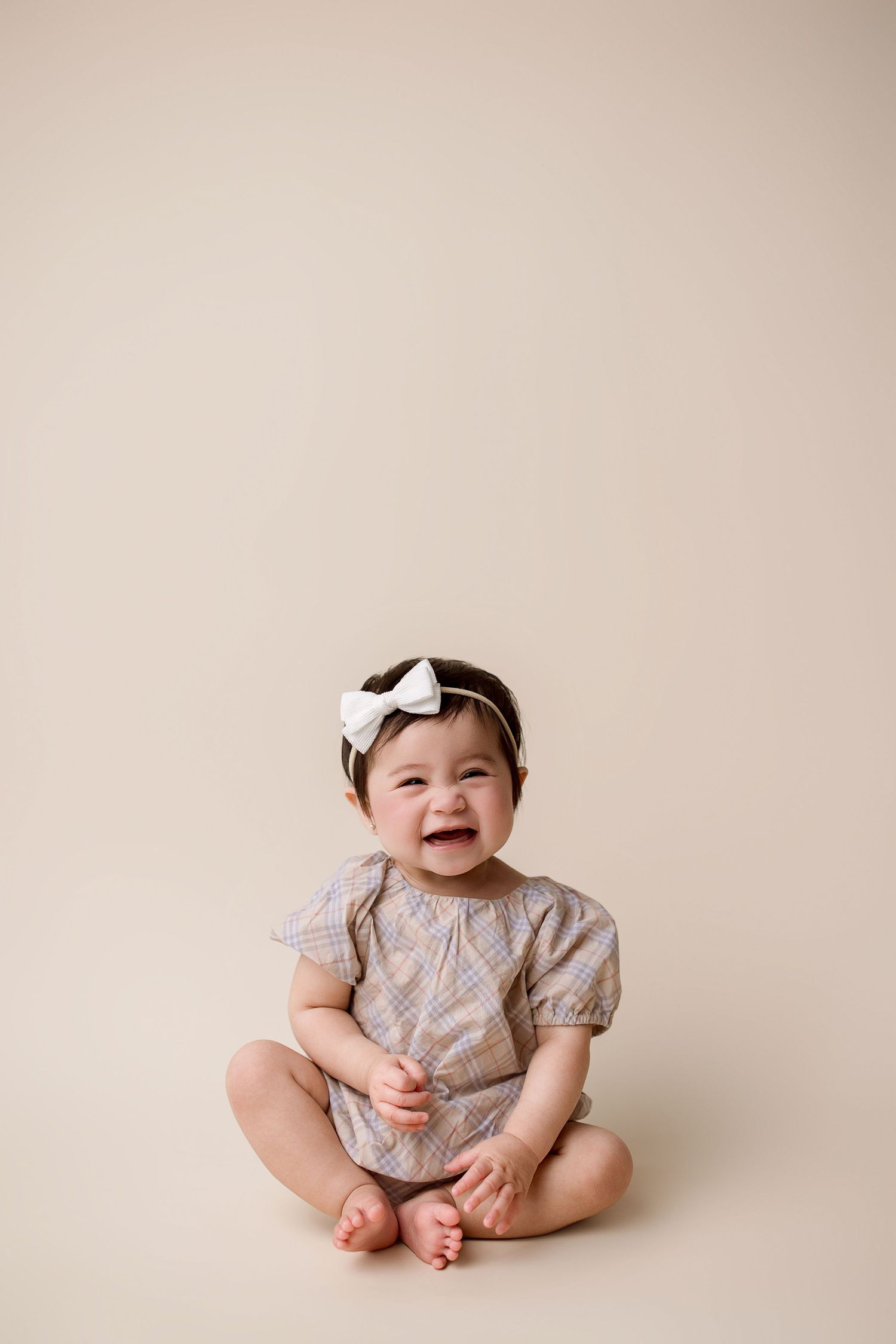 Baby in a floral dress sitting on a neutral background with a white bow accessory.