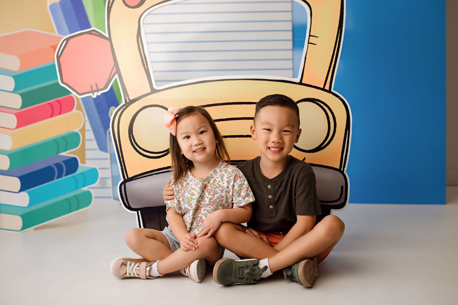 Two children sit cross-legged in front of colorful school bus themed backdrop.