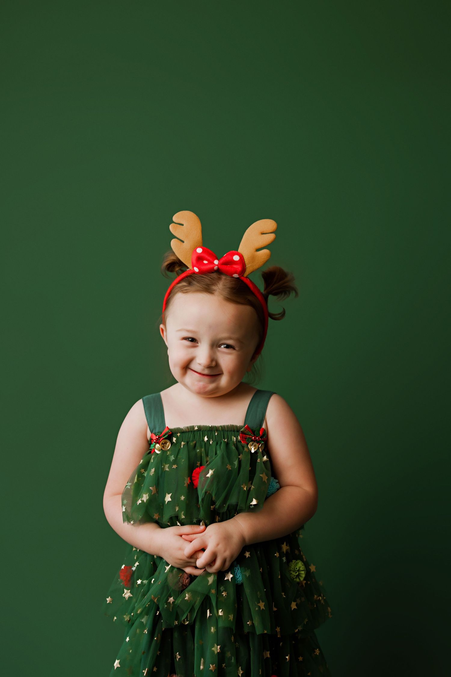 Child in green holiday dress wearing reindeer antlers smiles at camera against dark green backdrop.