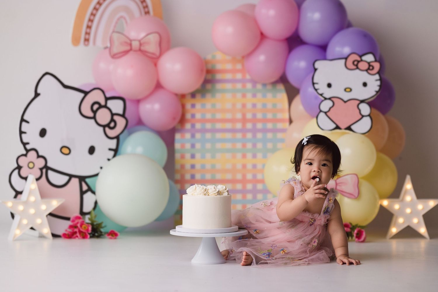 Baby girl in pink tutu sits before Hello Kitty backdrop with colorful balloon arch and white smash cake for first birthday.