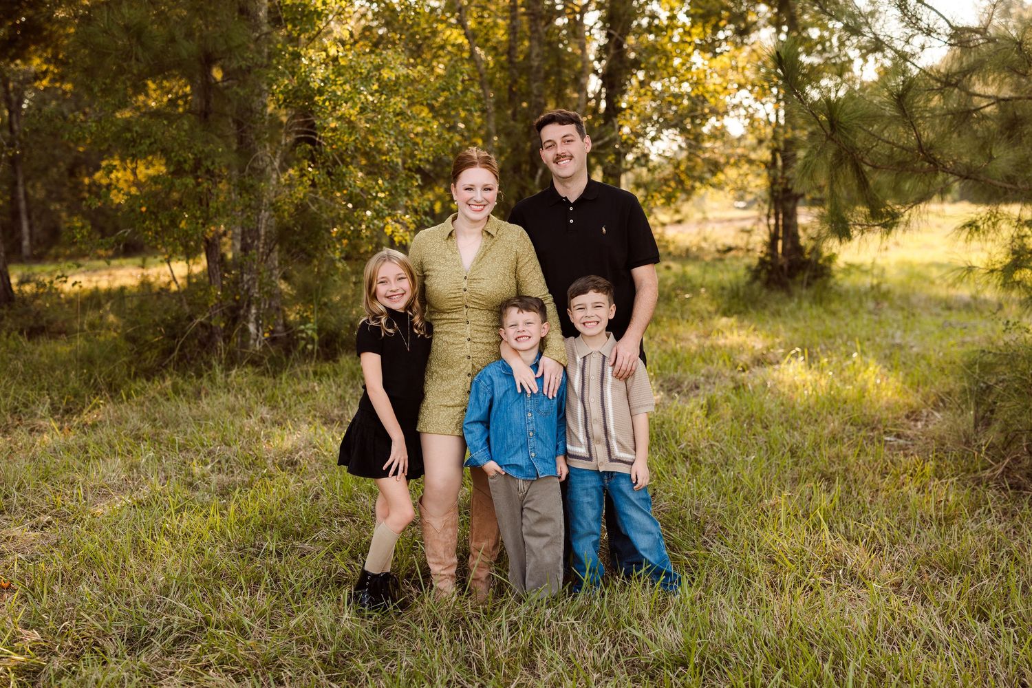 Family of five posing outdoors in a sunny grassy field with tall trees, two adults standing behind three smiling children.