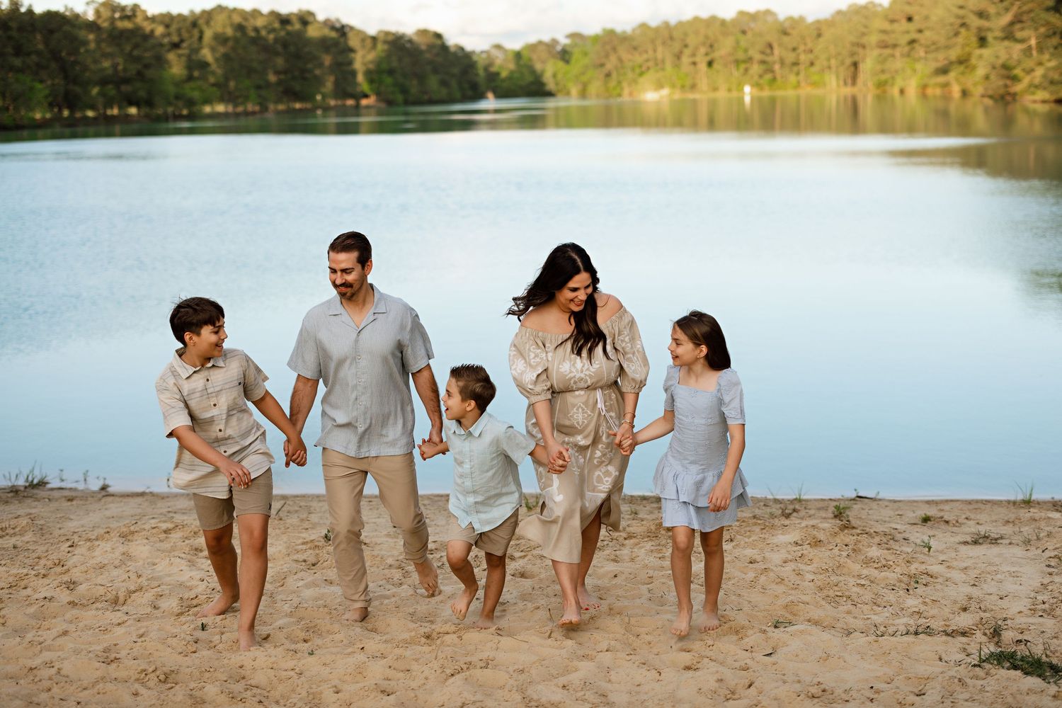 A family walks together along a lakeside beach during golden hour wearing matching neutral tones.