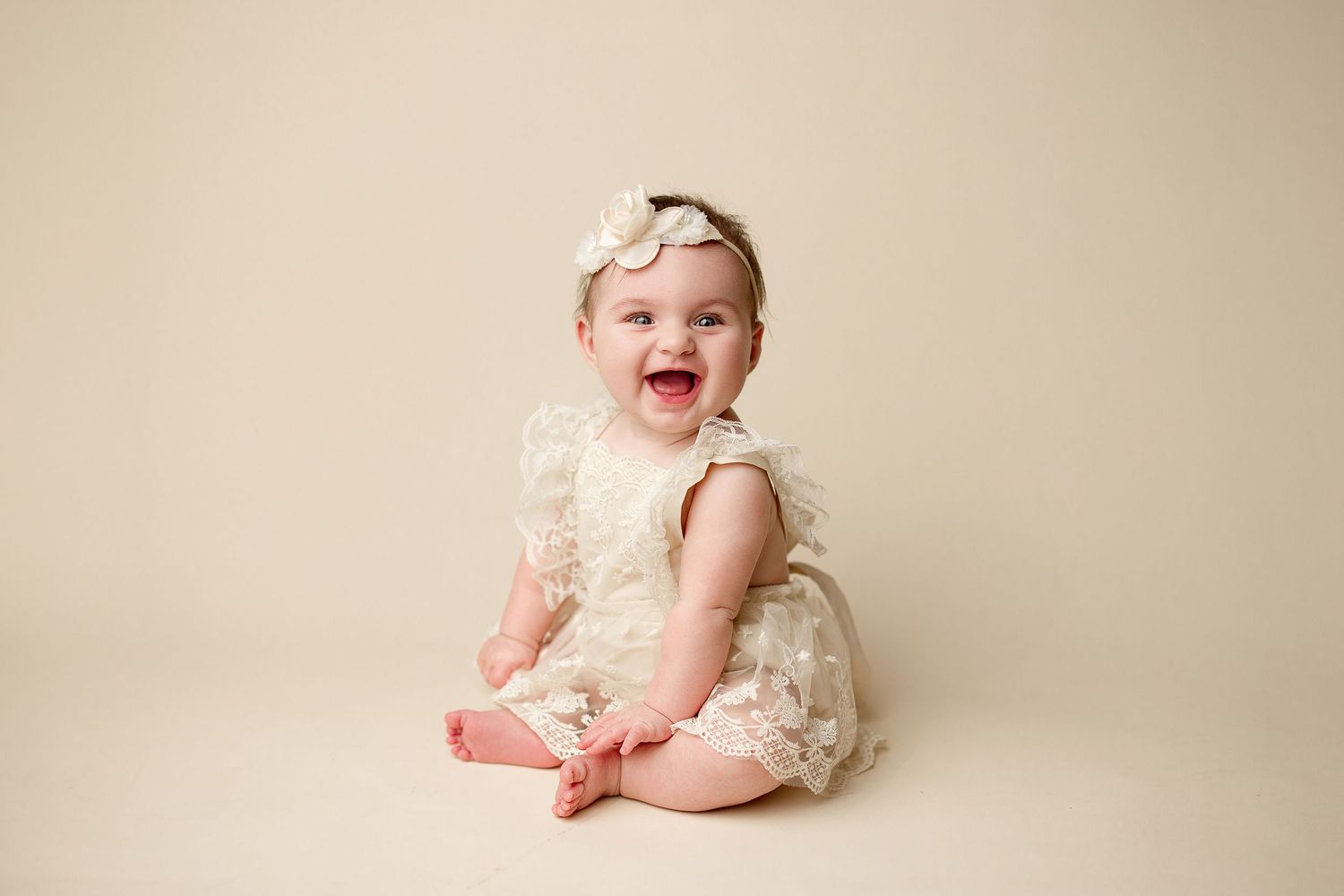 Baby in cream-colored lace dress sits and smiles on beige backdrop showing playful expressions in photo series.