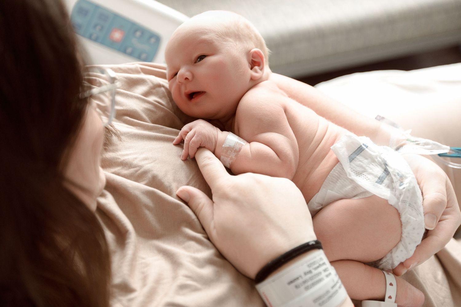 A peaceful newborn baby wearing a diaper rests on soft bedding while being gently touched by caring hands during Fresh 48 session.