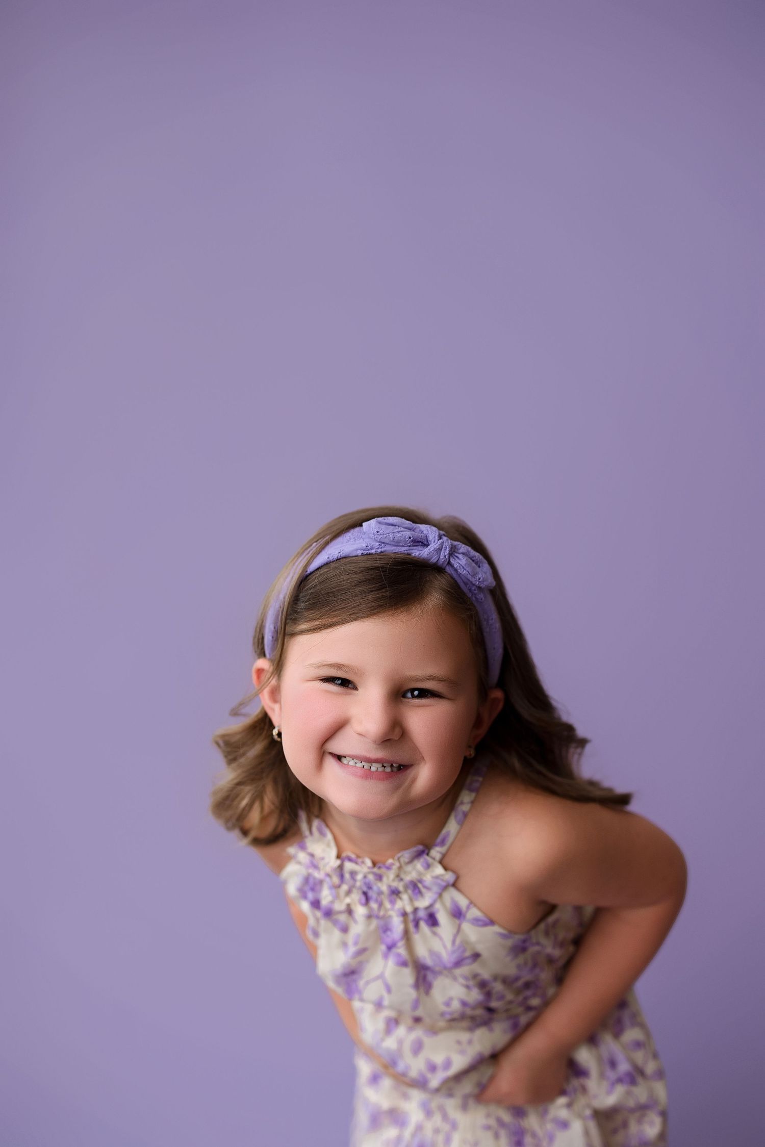 A child in a floral dress poses playfully against a purple background while smiling at the camera.