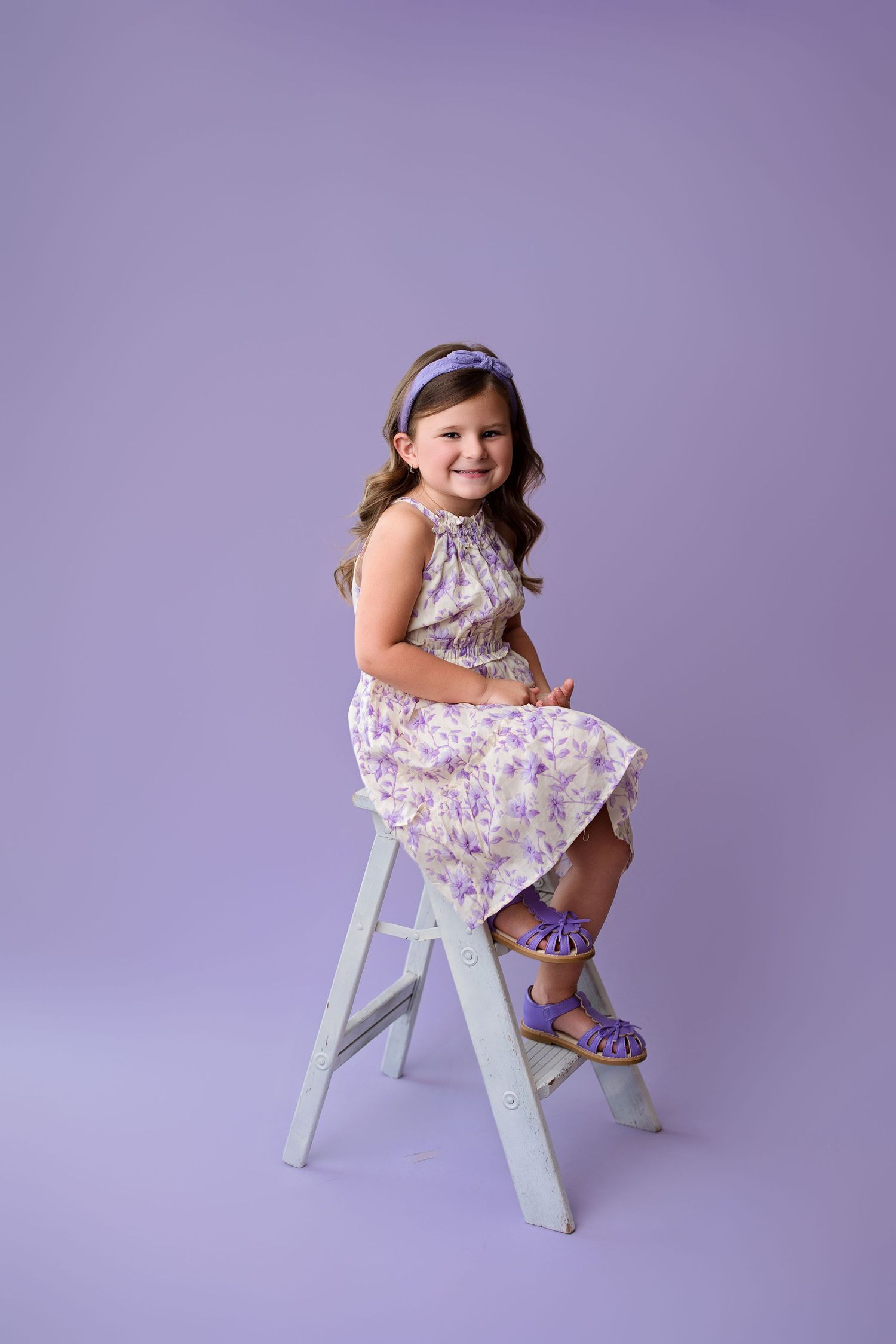 A floral dress-wearing child poses playfully on a white stool against a purple studio backdrop in a series of photos.