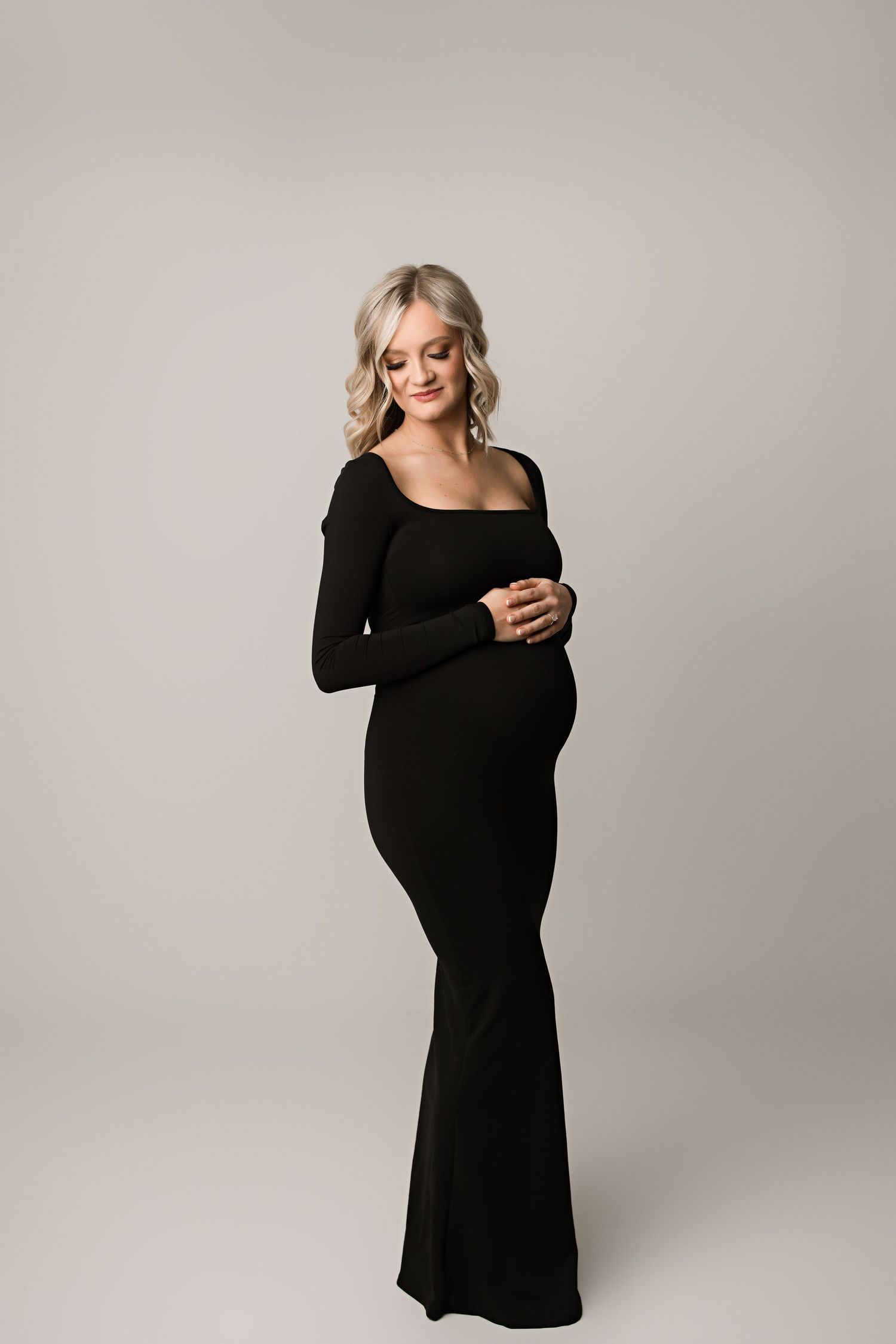 Elegant maternity portrait featuring a long black fitted dress against a neutral studio backdrop.