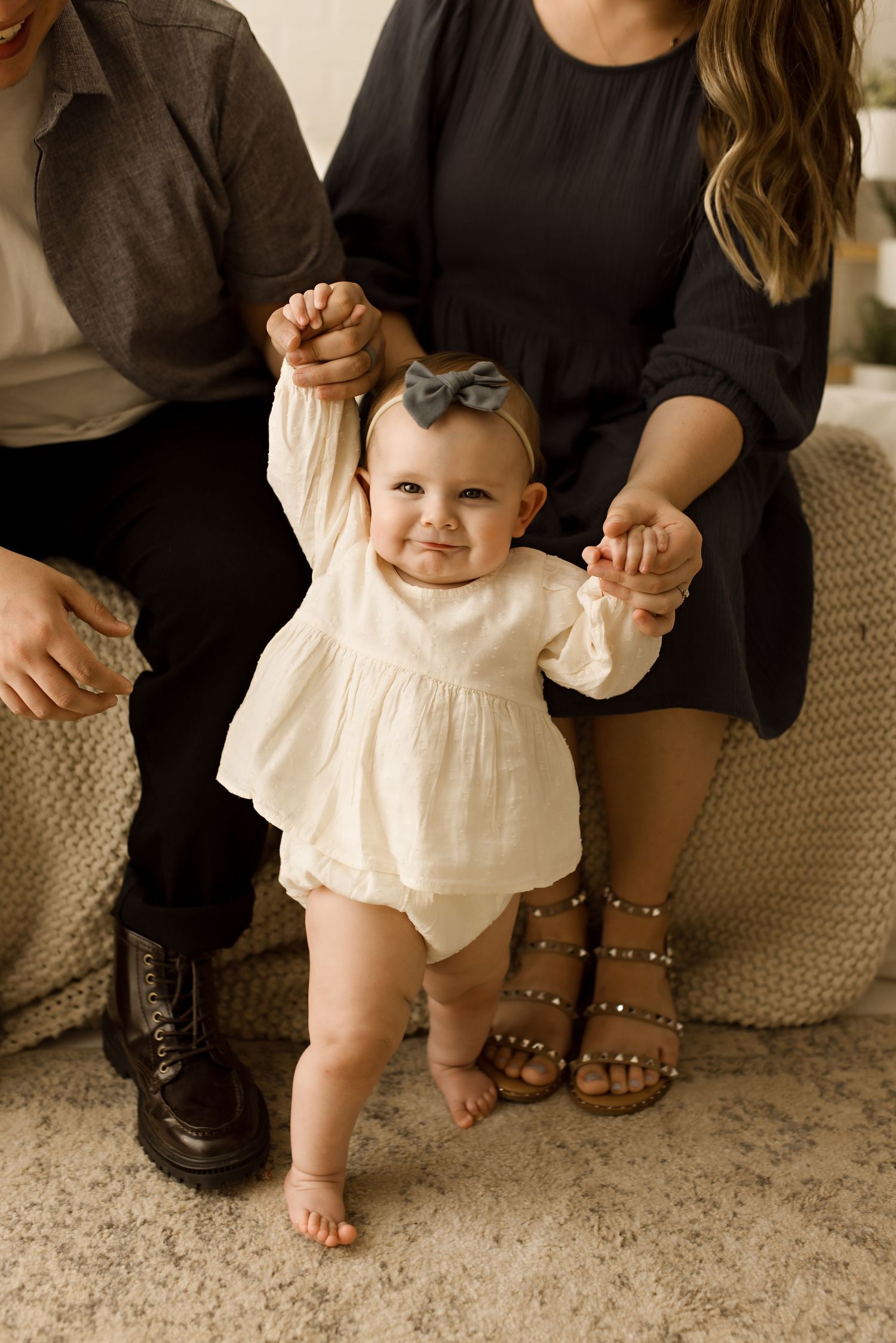 Baby taking first steps in cream dress while supported by parents.