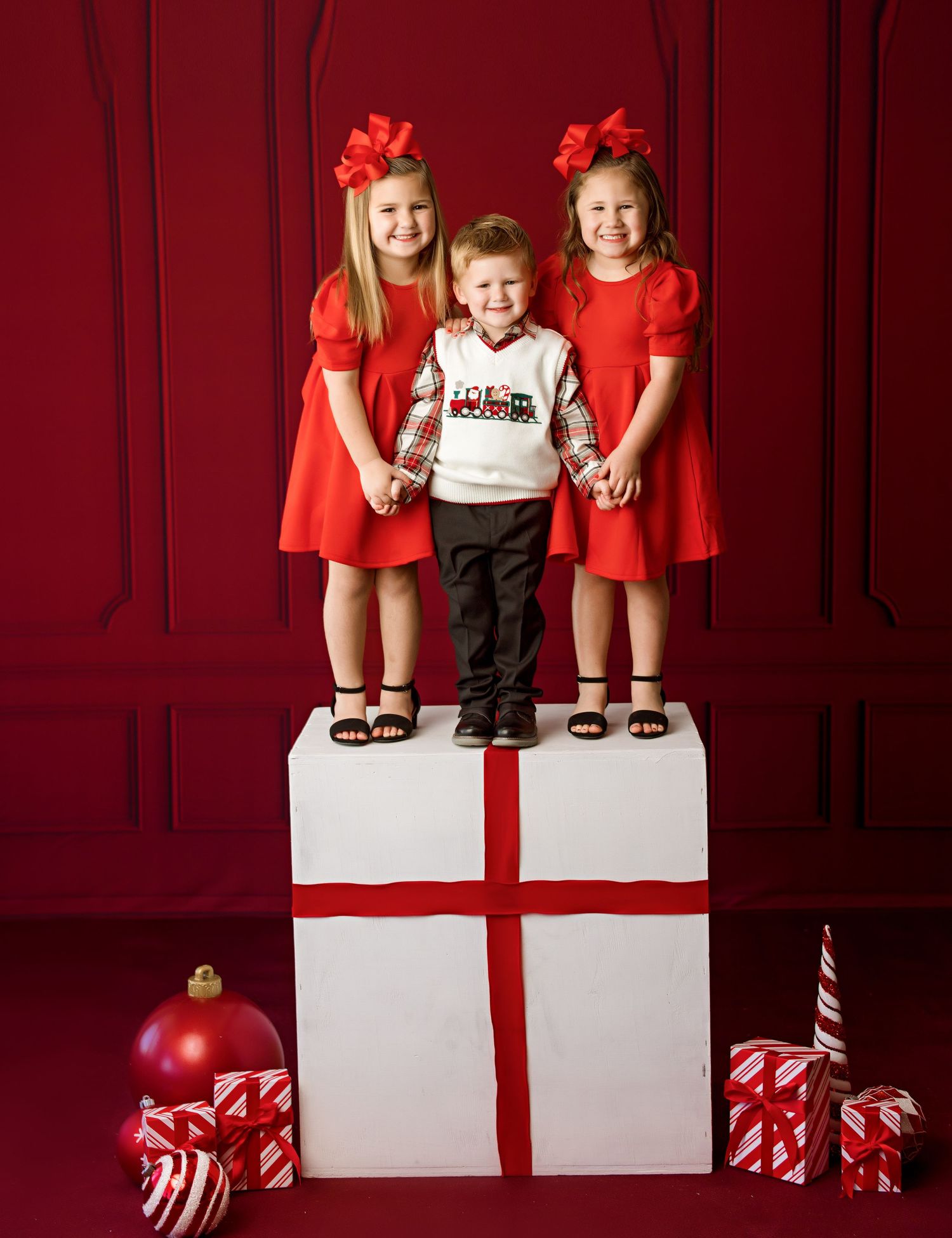 Three young children in red and white holiday outfits pose on a large gift box with Christmas decorations.