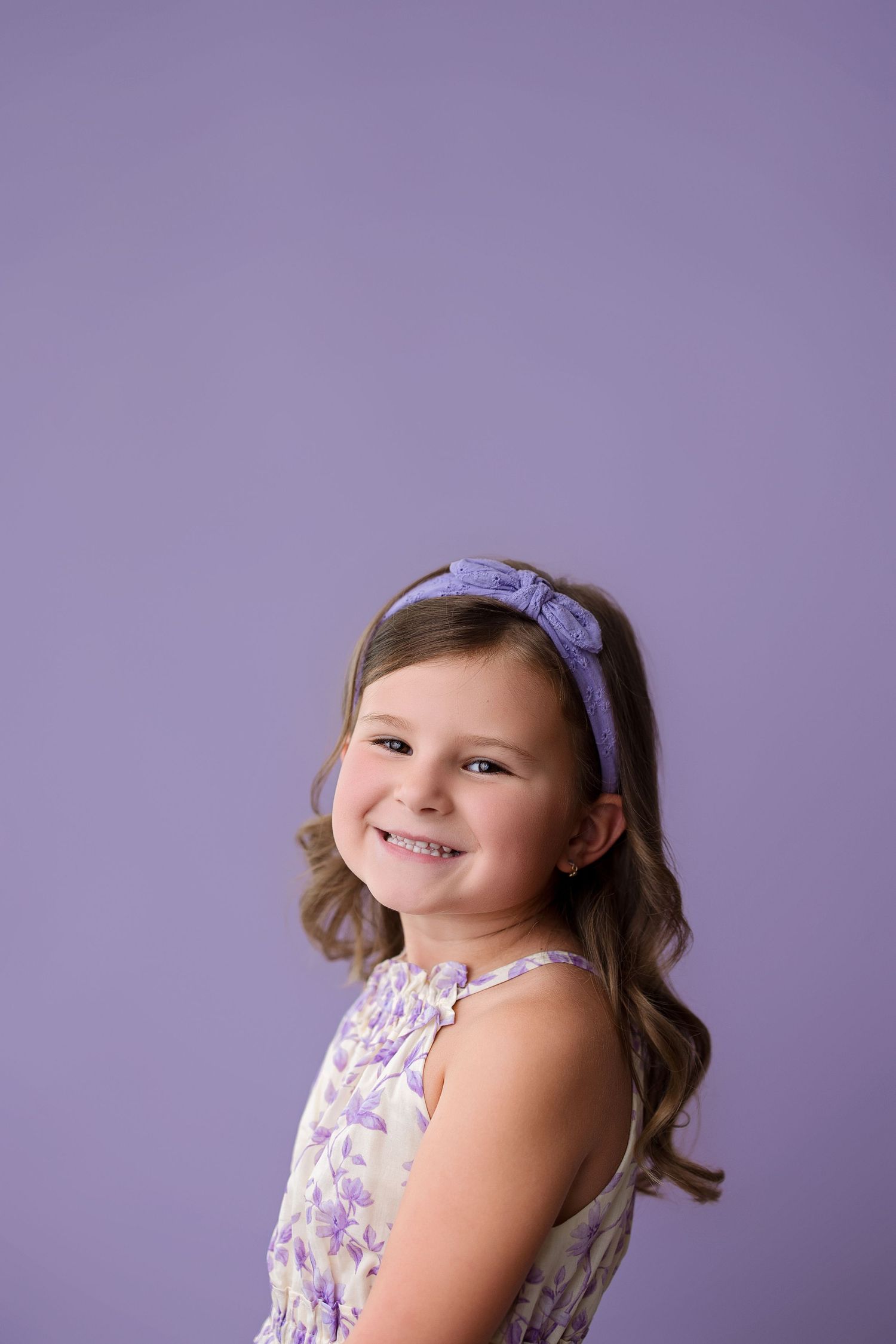 Young girl wearing purple floral dress and hair bow poses against lavender backdrop during portrait session.