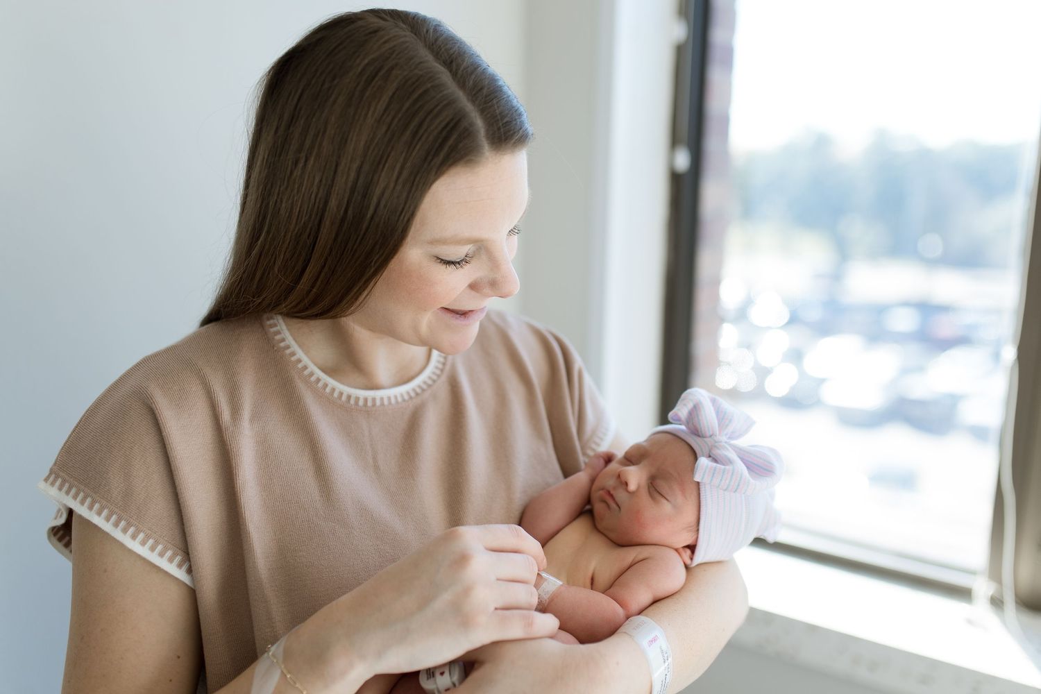 A mom in a beige top holds and gazes lovingly at a newborn baby near a bright window during Fresh 48 session in Beaumont, TX with Bree Elle.