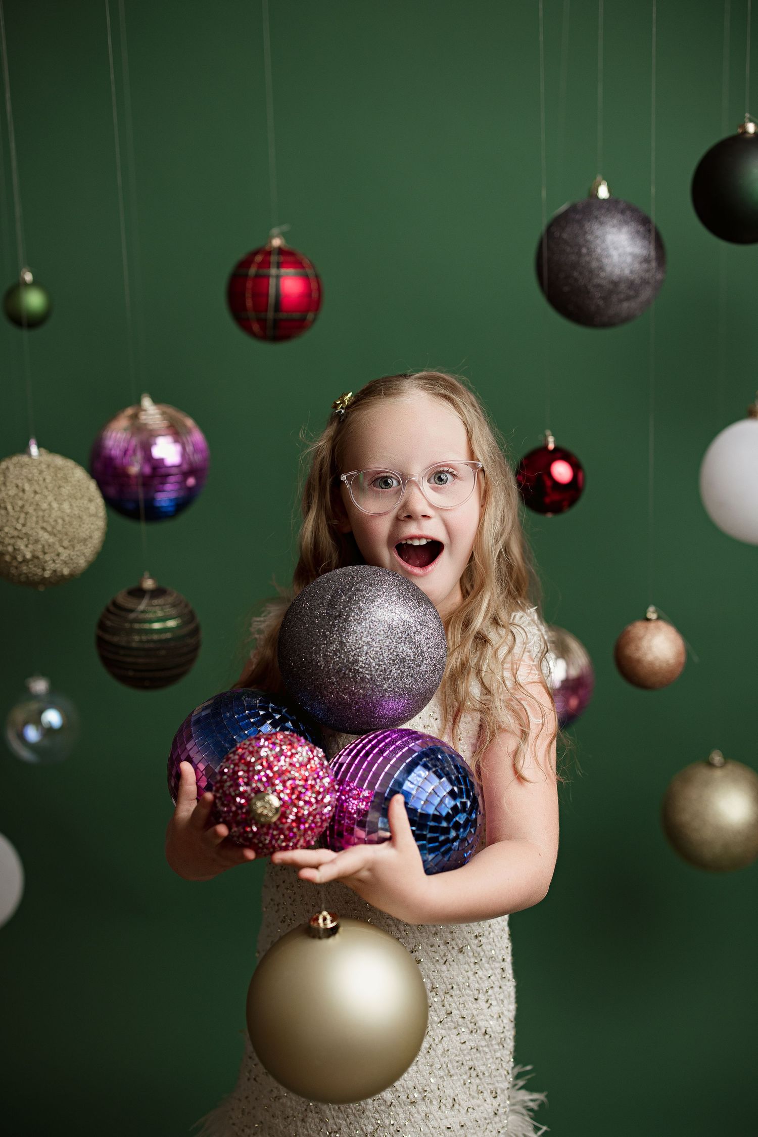 Colorful Christmas ornaments hang from above while a young child in white dress plays with festive decorations.