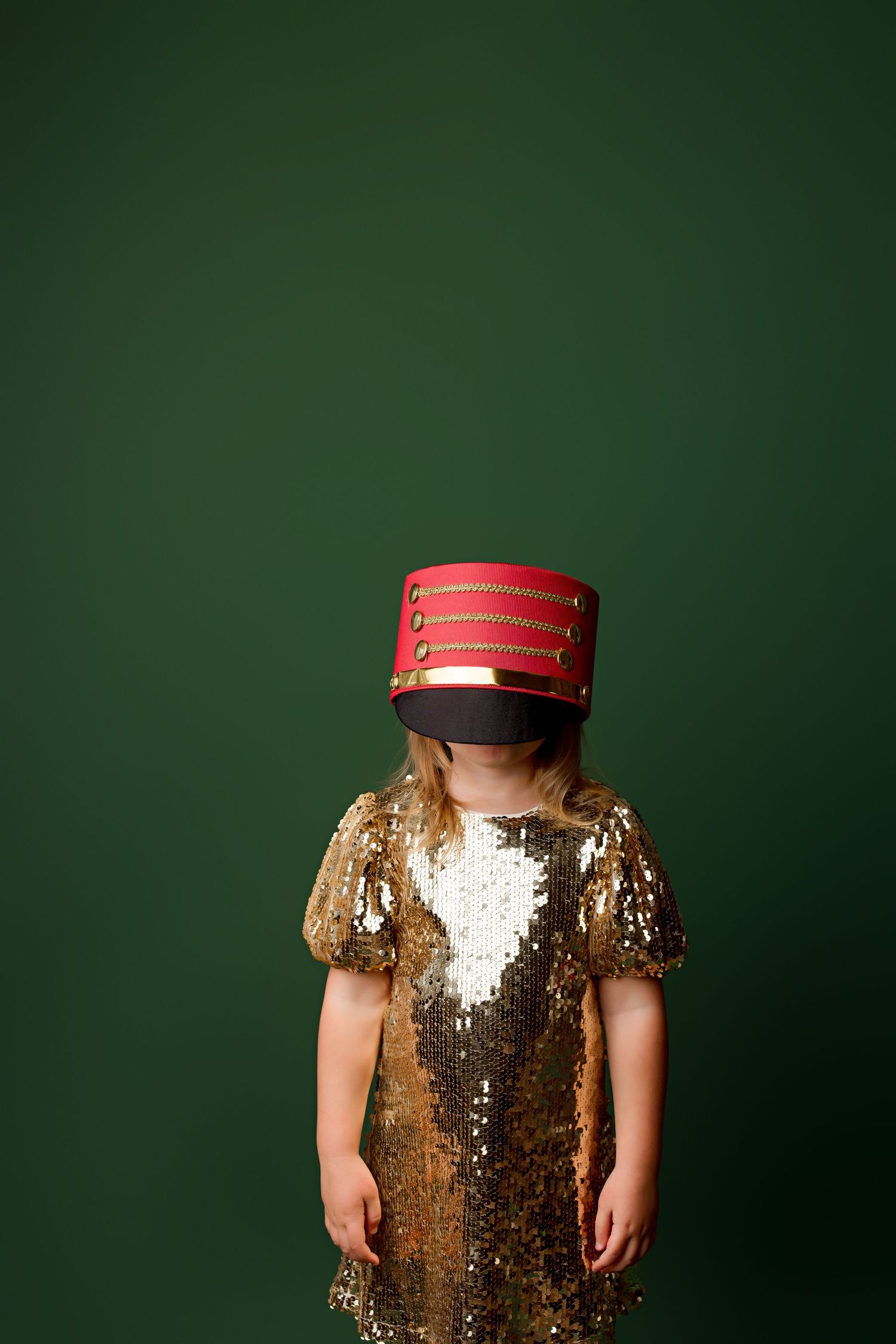Child wearing gold sequin dress and red marching band hat stands against green background.