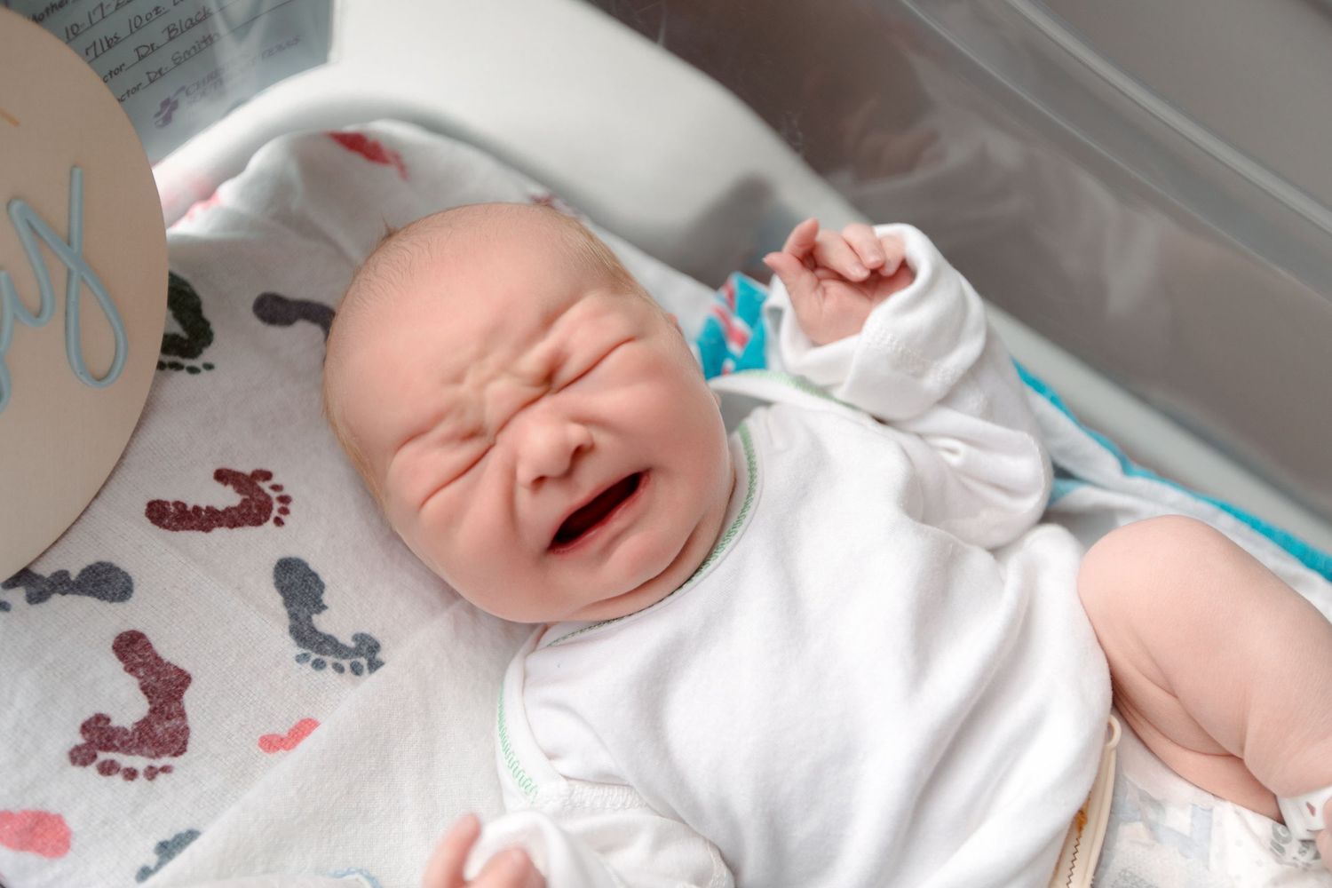 A newborn baby in white clothing cries while lying on patterned hospital bedding during Fresh 48 session in Beaumont hospital.