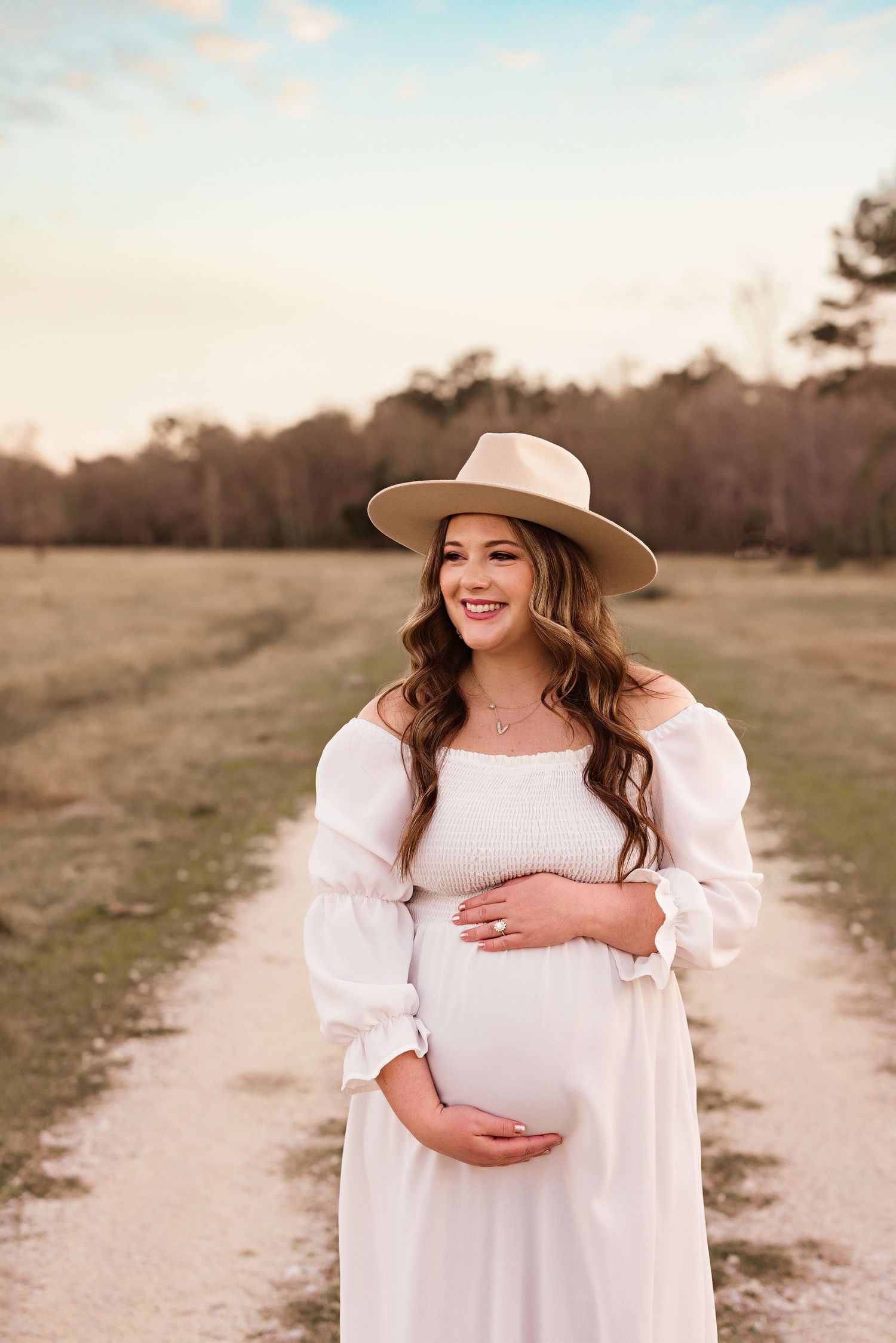 Pregnant woman in white dress and sun hat poses in a field during golden hour with Bree Elle Photography in Orange, TX.