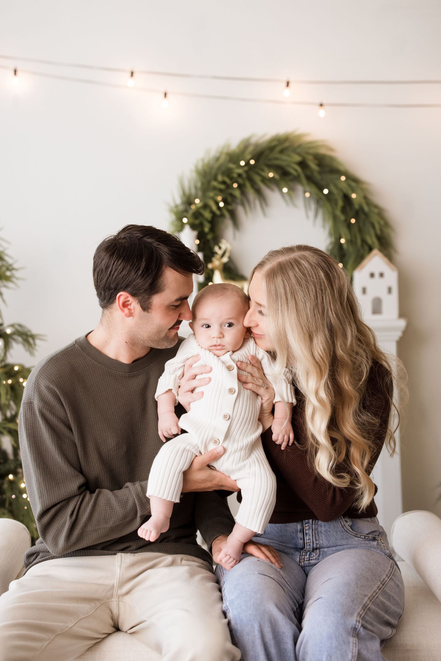 A family sits together in front of a holiday wreath and string lights for a cozy Christmas portrait.