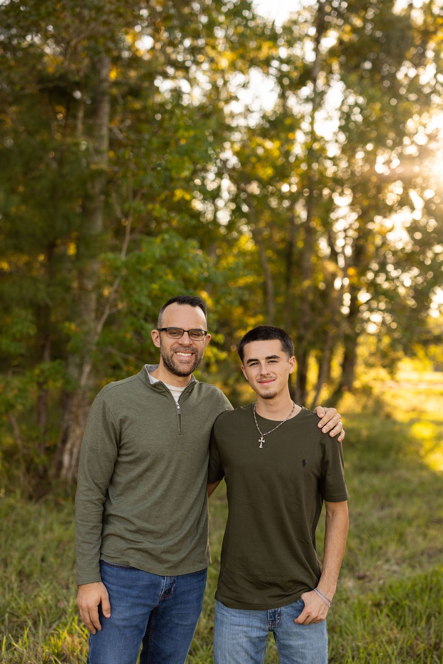 Two people stand together outdoors during golden hour with trees and natural lighting in the background.