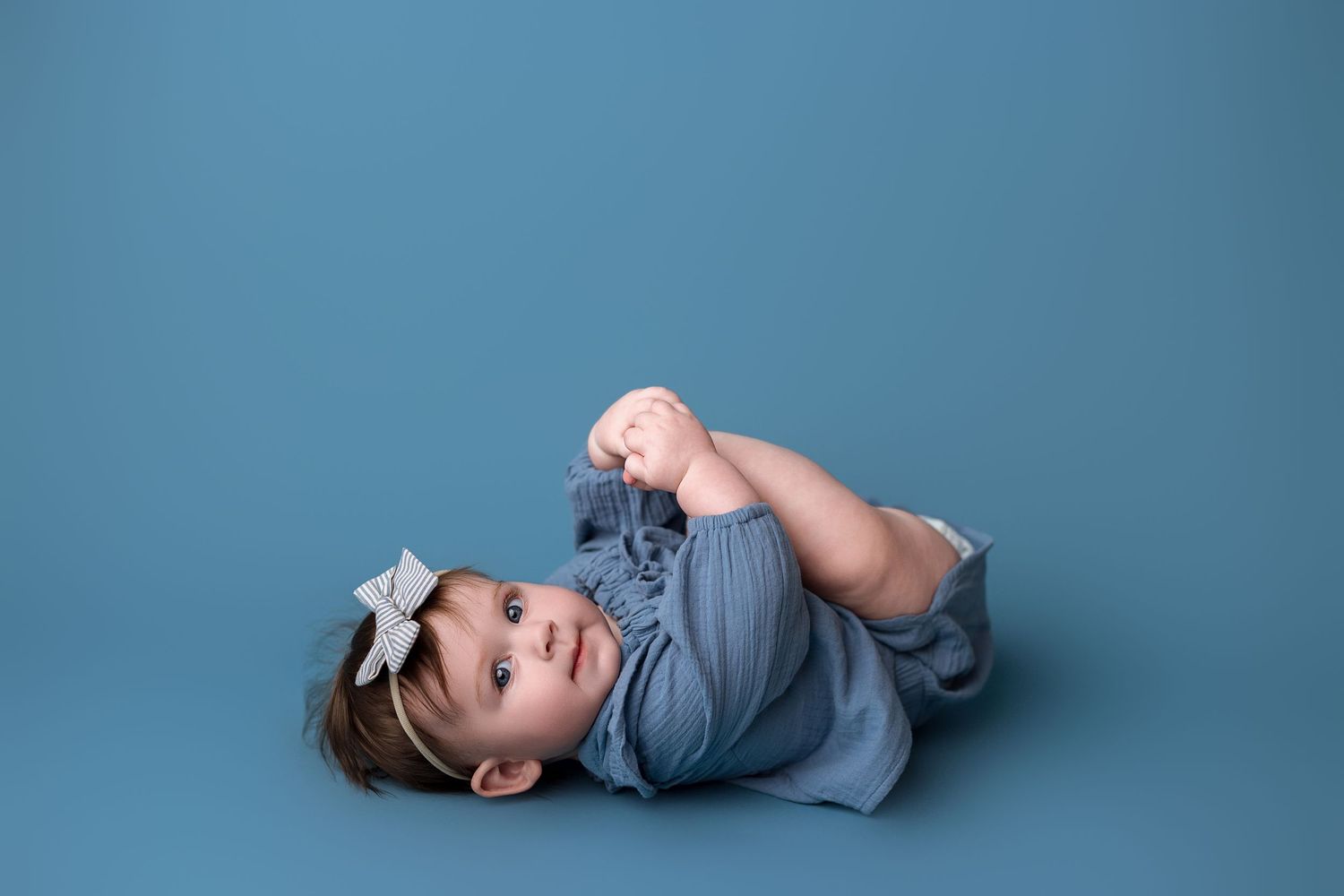 A sleeping baby in a grey onesie lies peacefully on a blue backdrop during a professional newborn photo session.